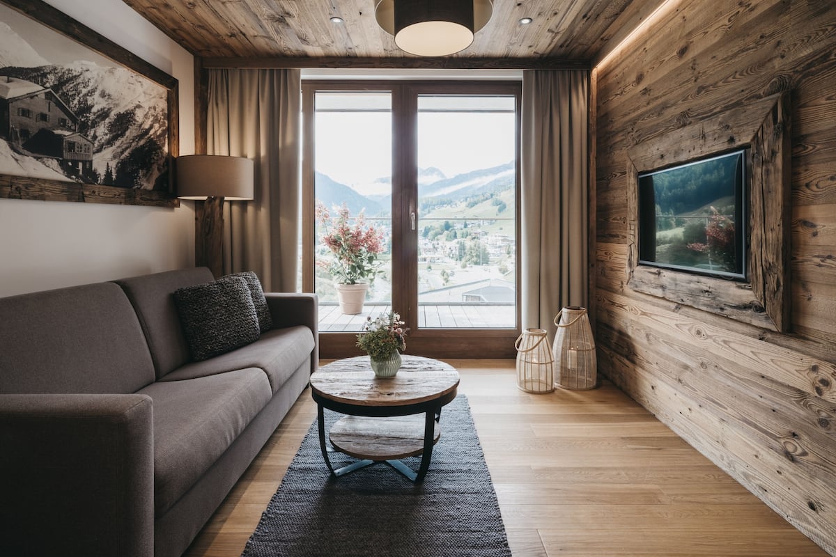 A cozy room with a wooden ceiling, a large window with curtains, a sofa, a small round table with a vase, and a framed picture on the wall.