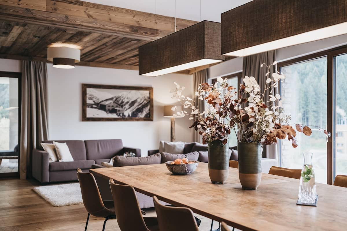 A modern living room with a wooden dining table, comfortable seating, a large window, and a decorative vase with flowers.