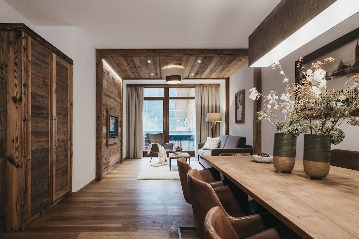 A modern interior with a wooden floor, a dining area with a long table and chairs, a sofa, and a large window with a view of the outdoors.
