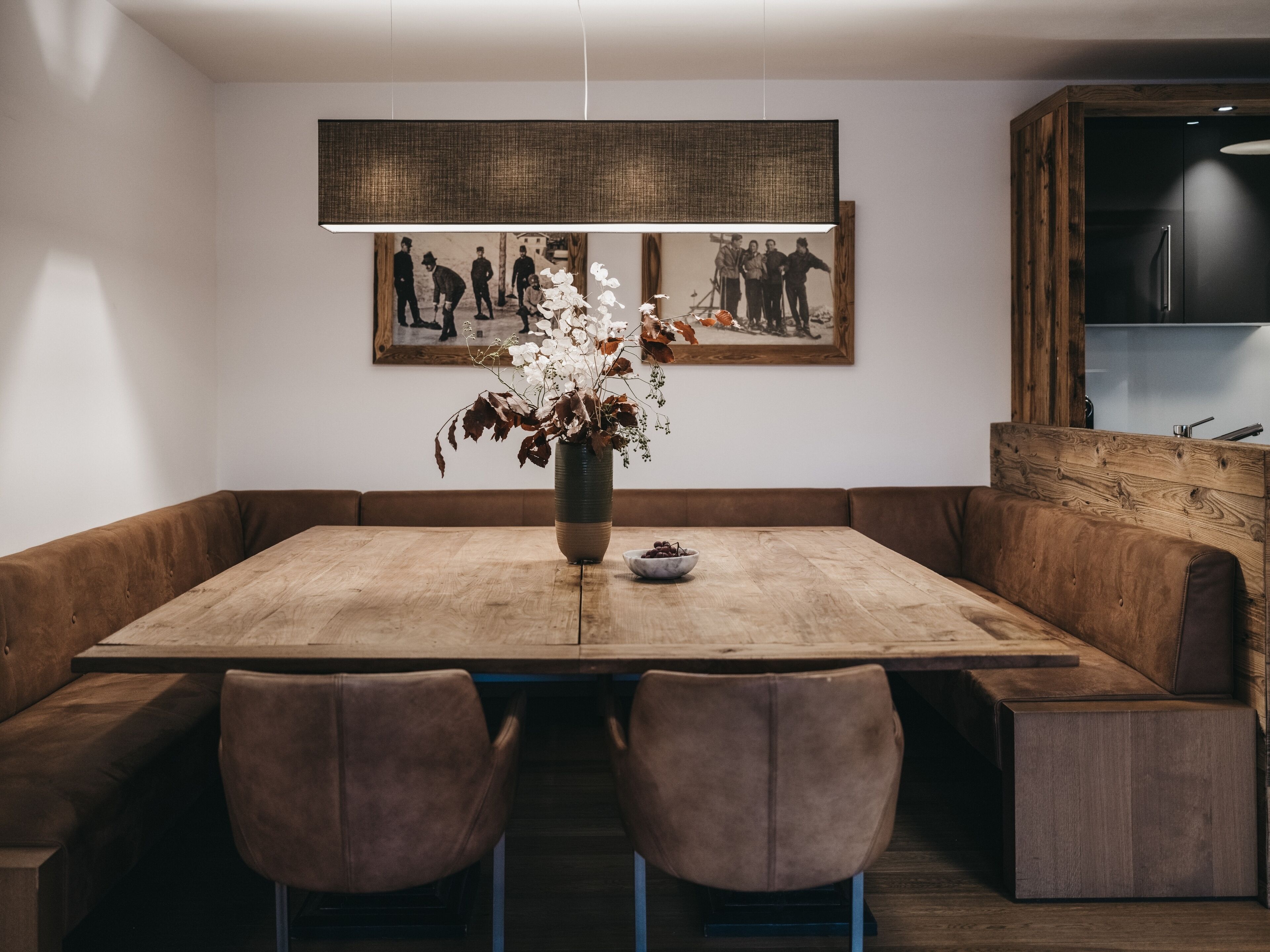 Un comedor con mesa de madera, bancos tapizados en marrón y un jarrón con flores.