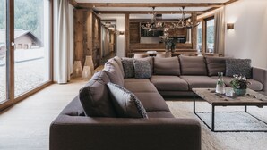 Uma sala de estar moderna com um amplo sofá modular, uma mesa de centro e vista para uma paisagem montanhosa através de grandes janelas.