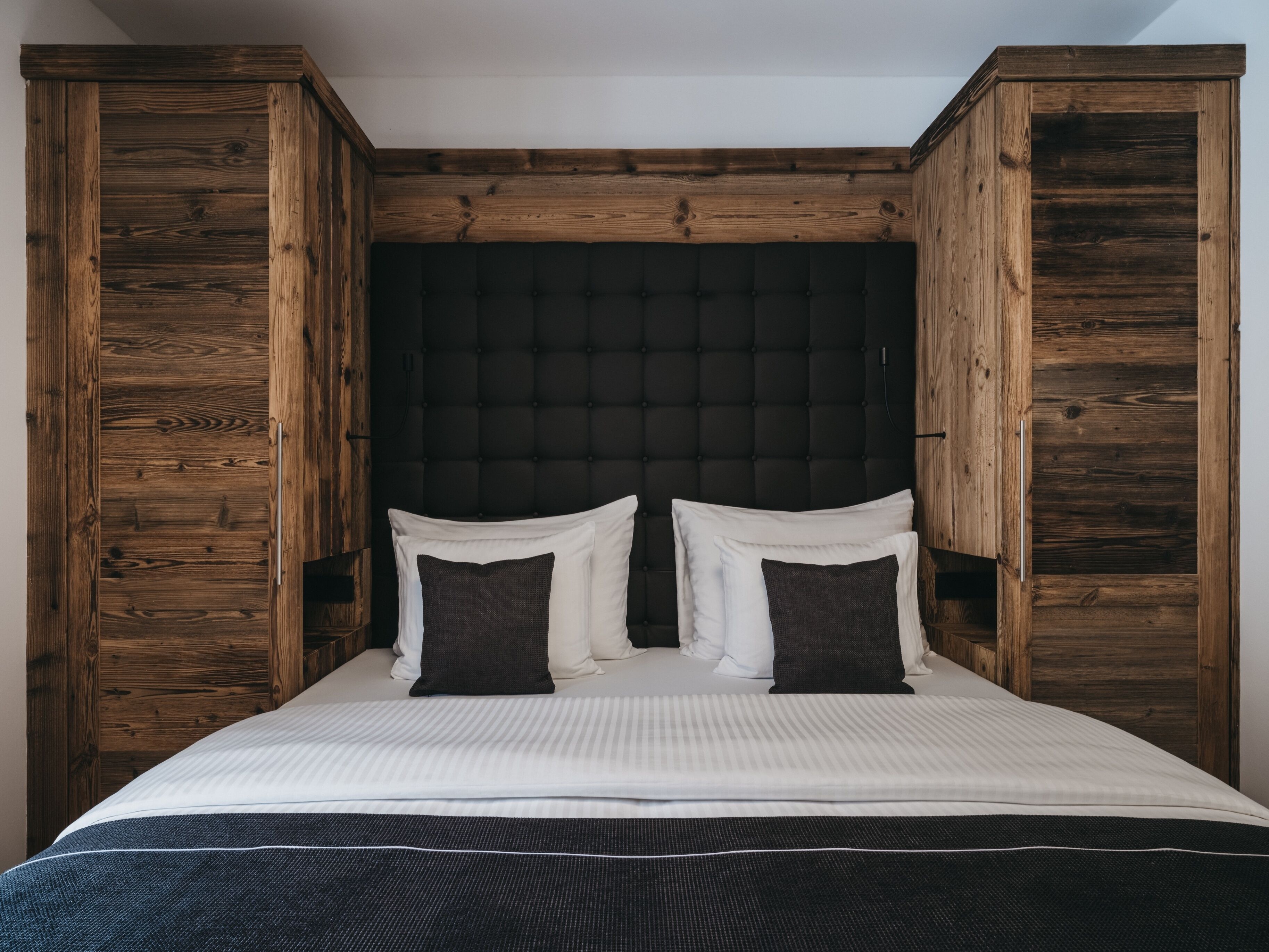 Uma cama com cabeceira escura, ladeada por painéis de madeira, num quarto com paredes brancas.