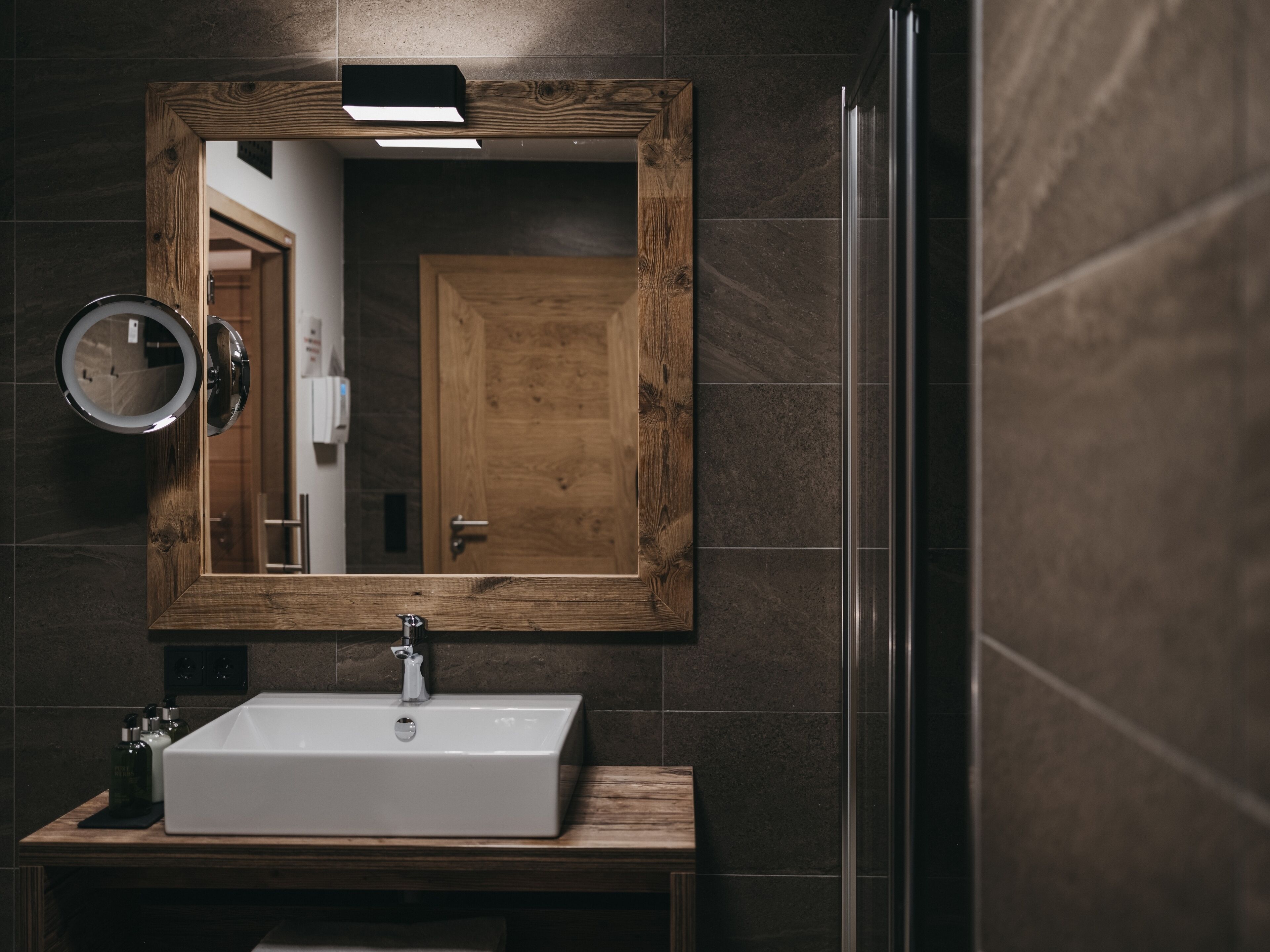 Un baño con un espejo de marco de madera, un lavamanos blanco y una mampara de vidrio para la ducha.