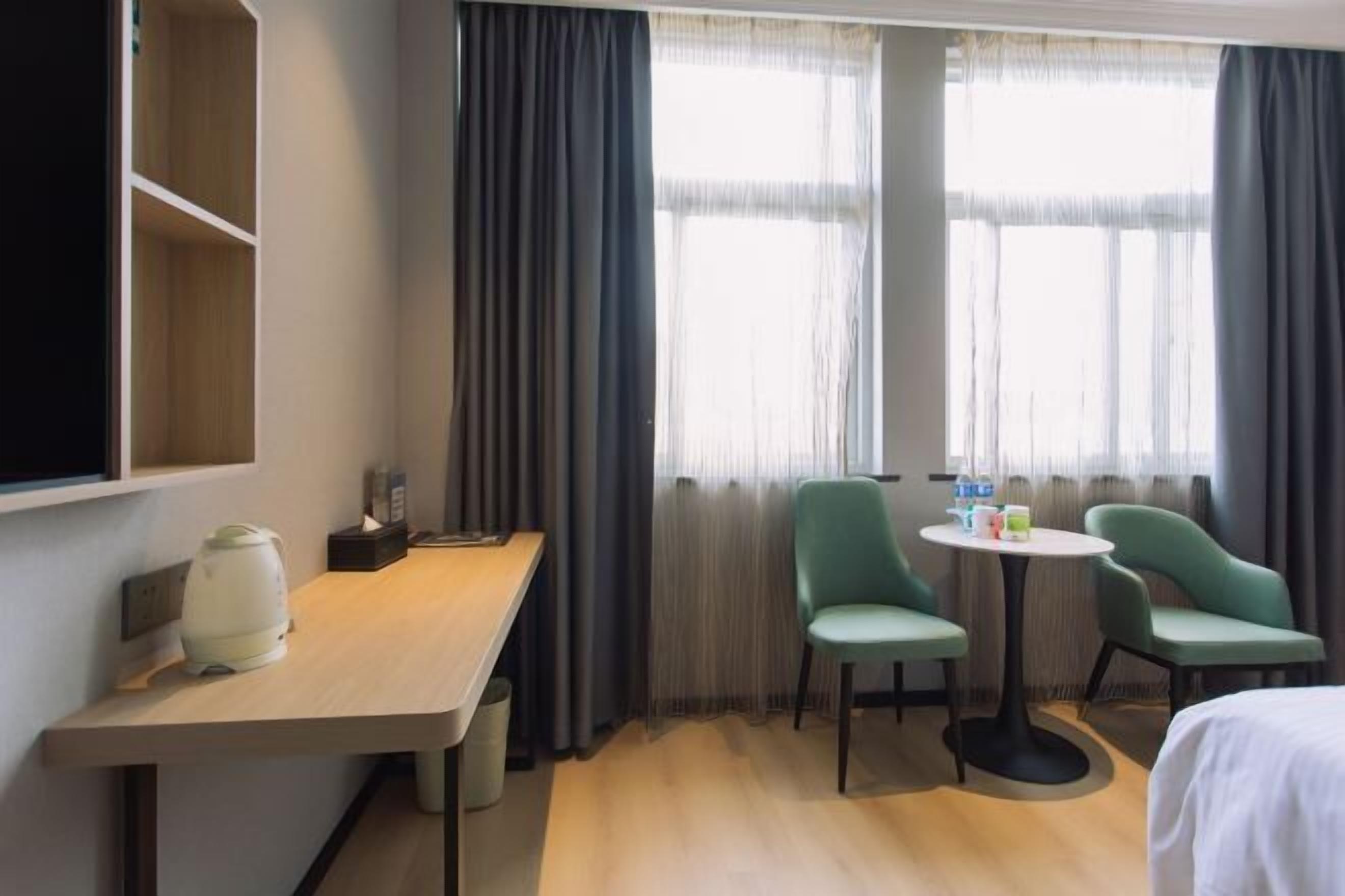 Habitación de hotel con un escritorio de madera, dos sillas verdes, una mesita con bebidas y una cama.