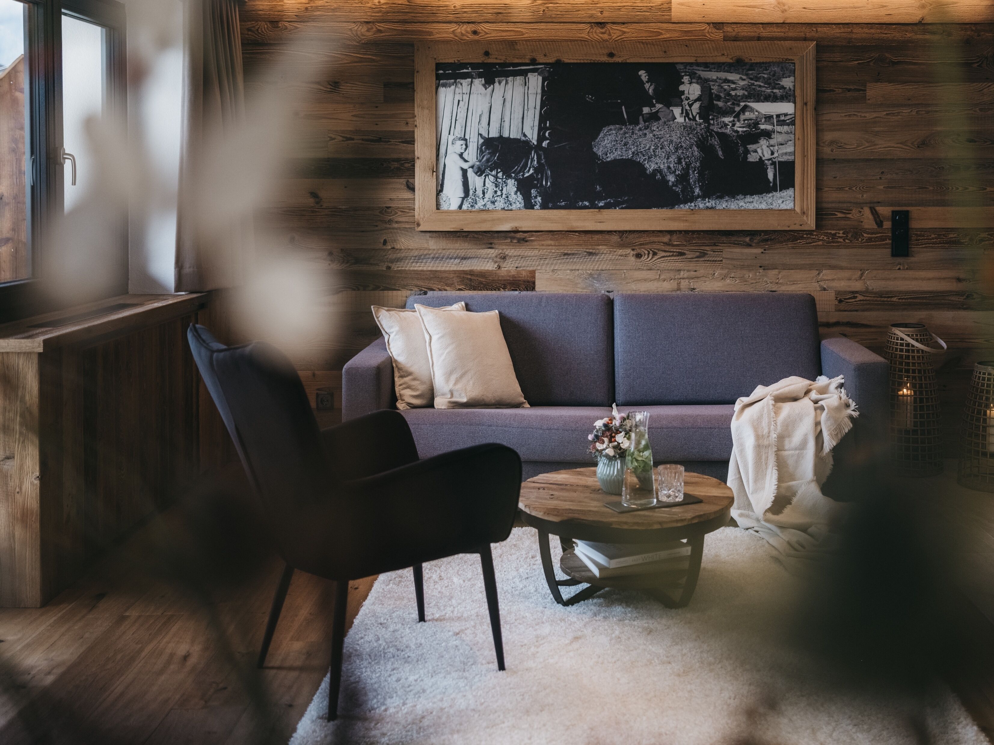 Een gezellige woonkamer met een houten wand, een grijze bank, een donkerblauwe fauteuil, een ronde houten salontafel, en een zwart-wit foto in een lijst aan de muur.