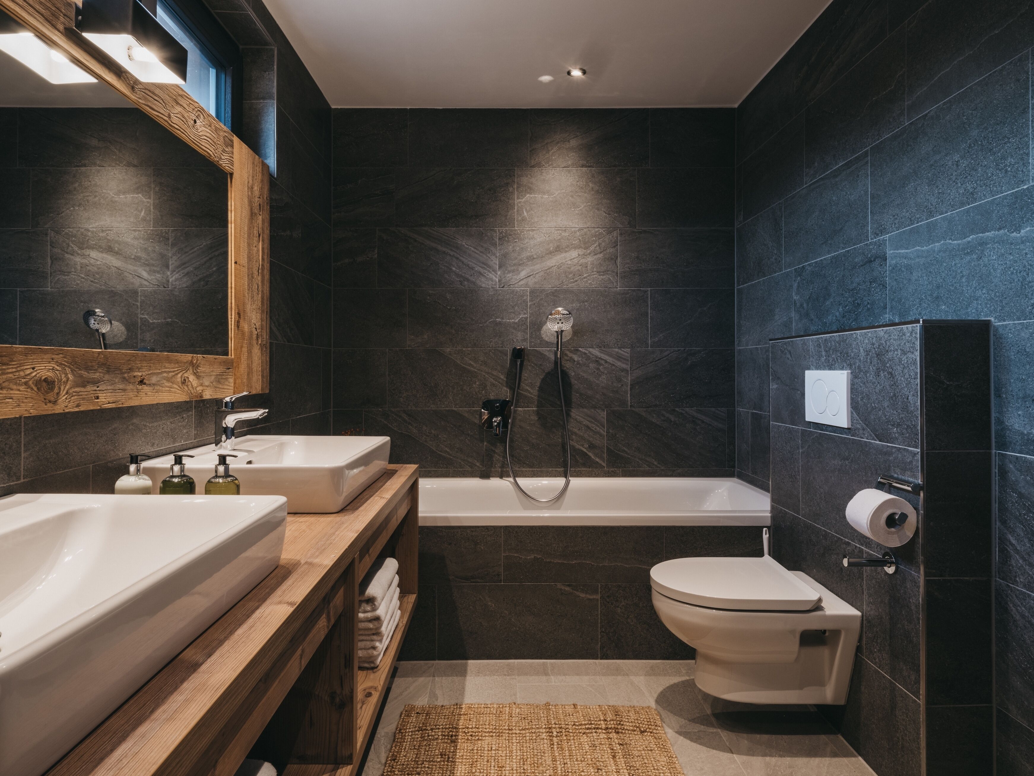 A modern bathroom with dark stone tiles, a wooden vanity, a bathtub, and a toilet.