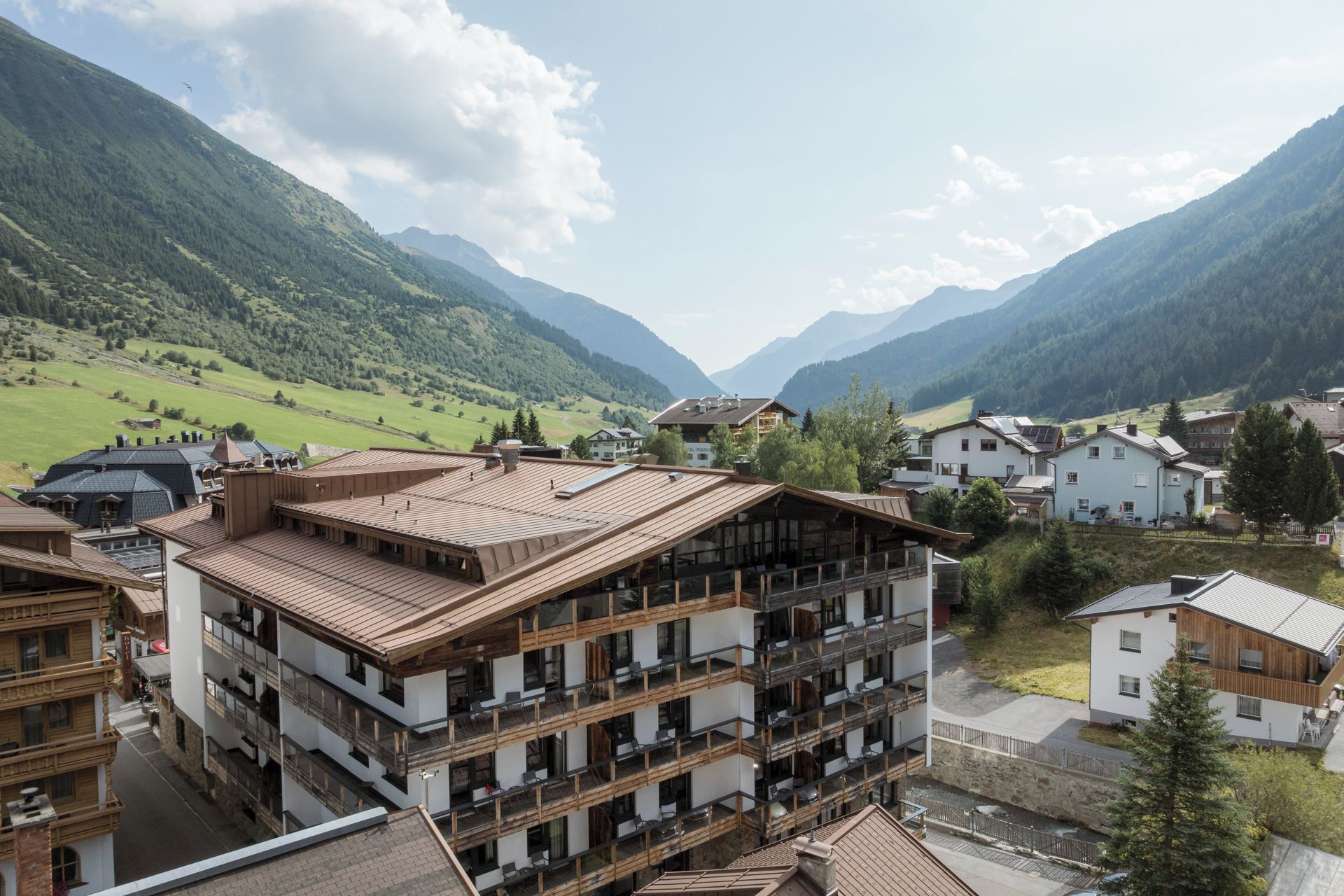 A multi-story building with balconies, set in a mountainous area with green valleys and houses.