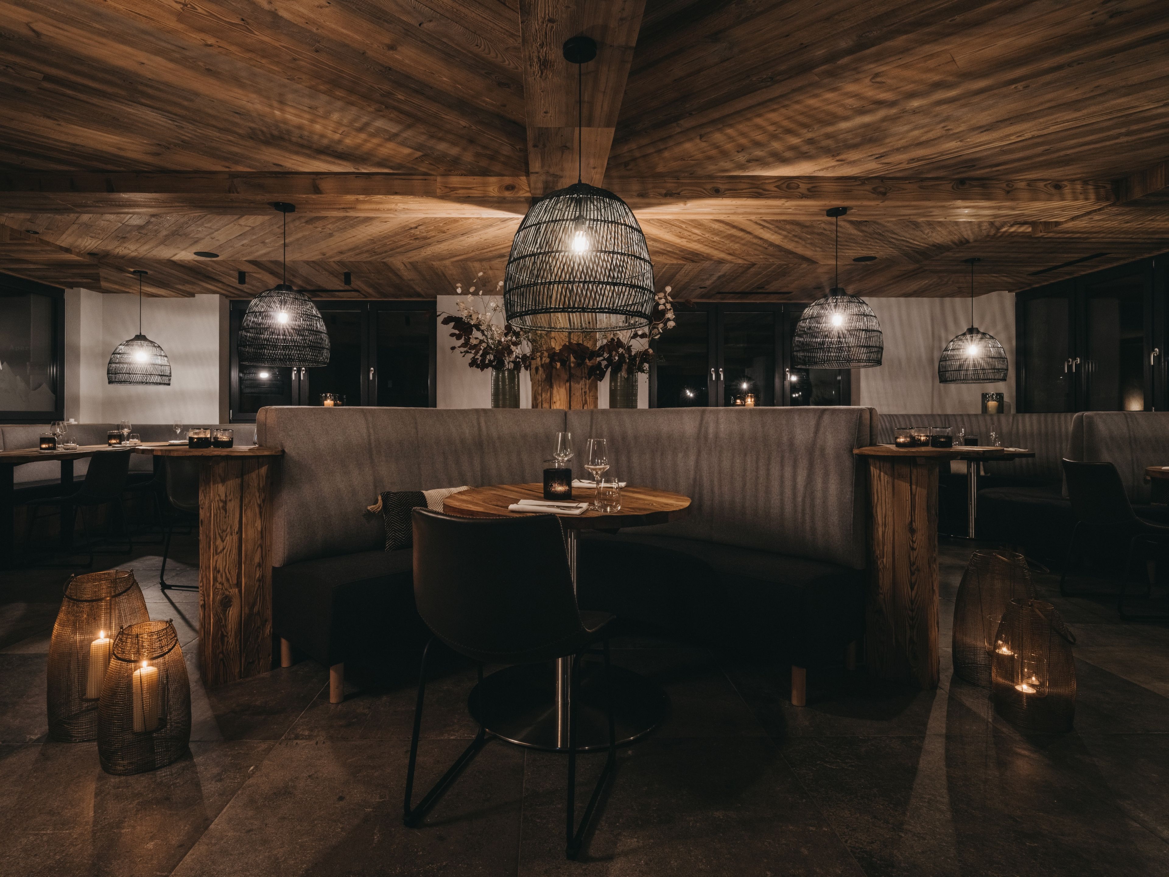 A dimly lit restaurant with wooden ceiling and walls, featuring round tables, cushioned seating, and hanging pendant lights.