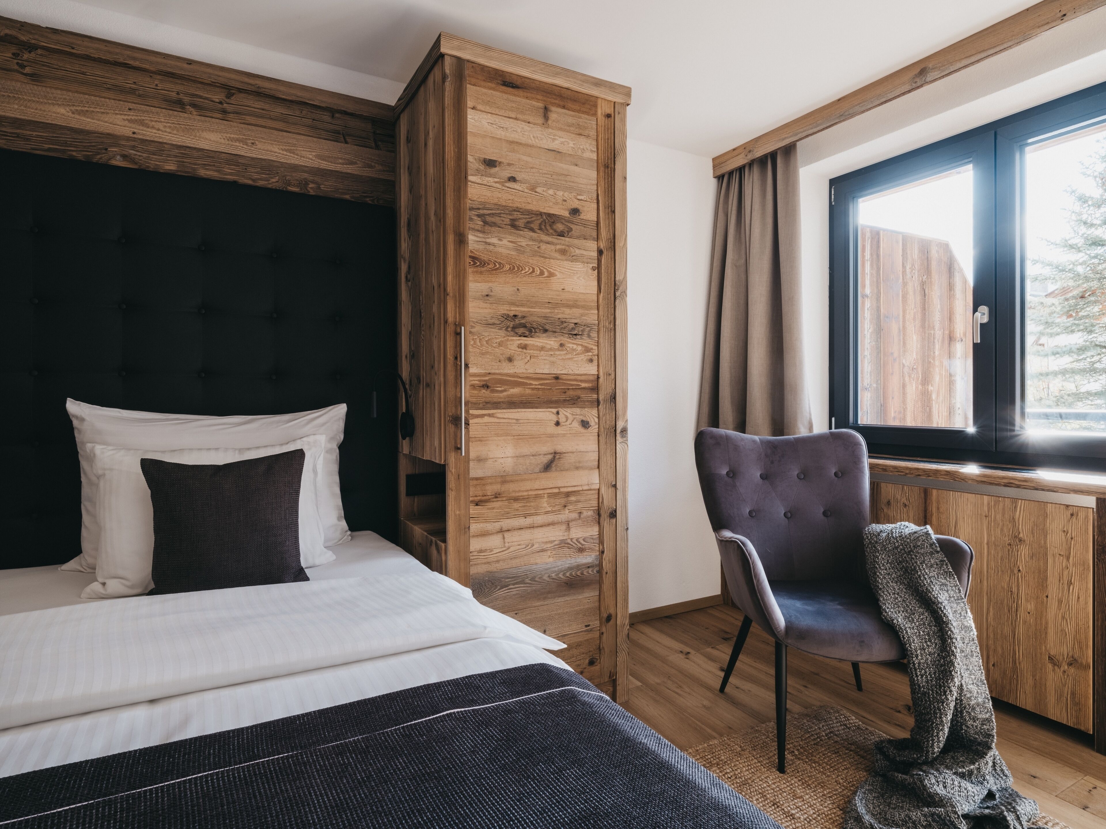 Une chambre à coucher avec un lit, une armoire en bois, une chaise et une fenêtre avec des rideaux.