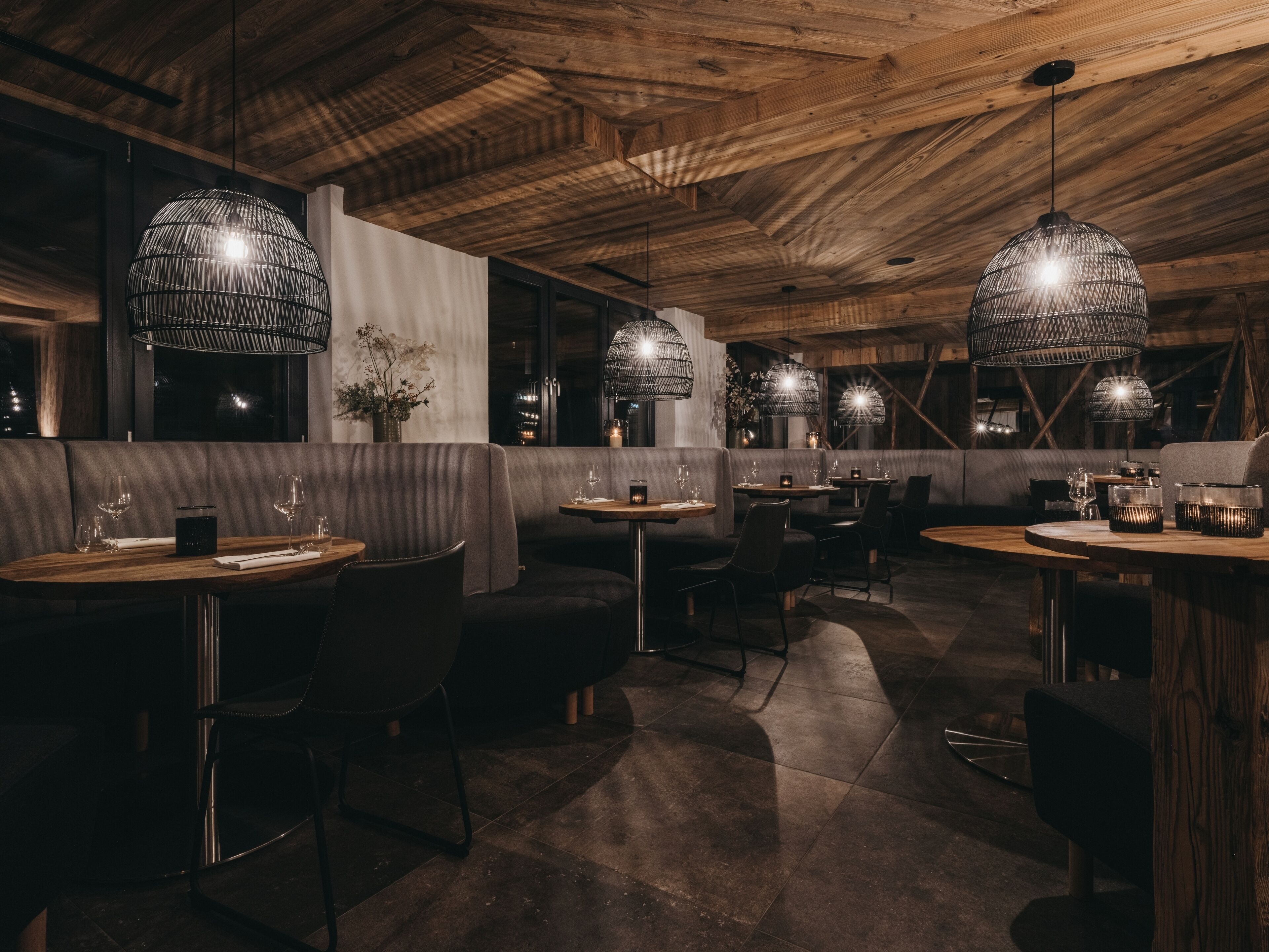 Interior de restaurante con vigas de madera en el techo, mesas redondas y sillas negras.