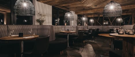 Interior de restaurante con vigas de madera en el techo, mesas redondas y sillas negras.