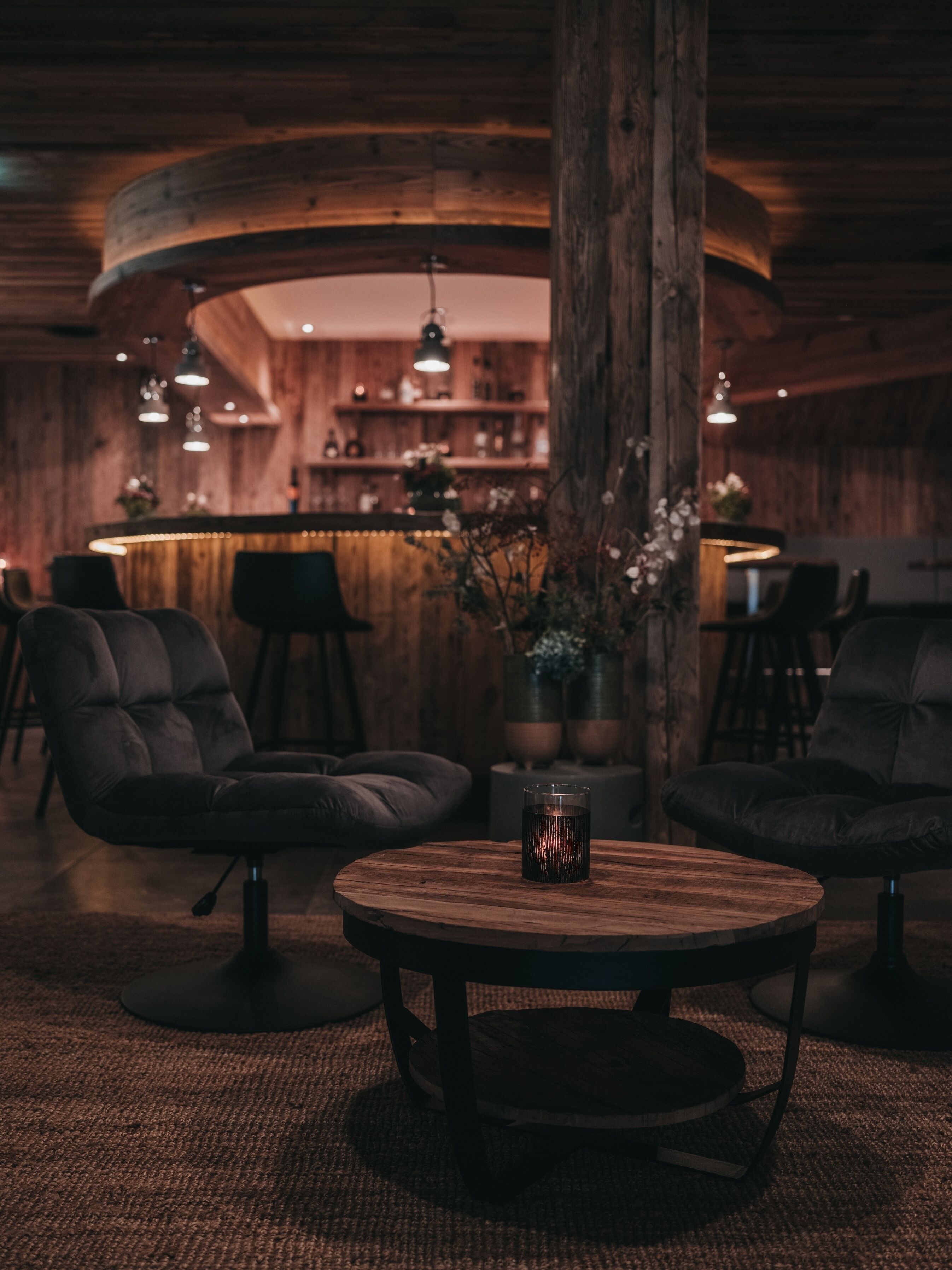 Une pièce chaleureuse et faiblement éclairée, avec un comptoir en bois, deux fauteuils en cuir et une table ronde en bois sur laquelle est posée une bougie.