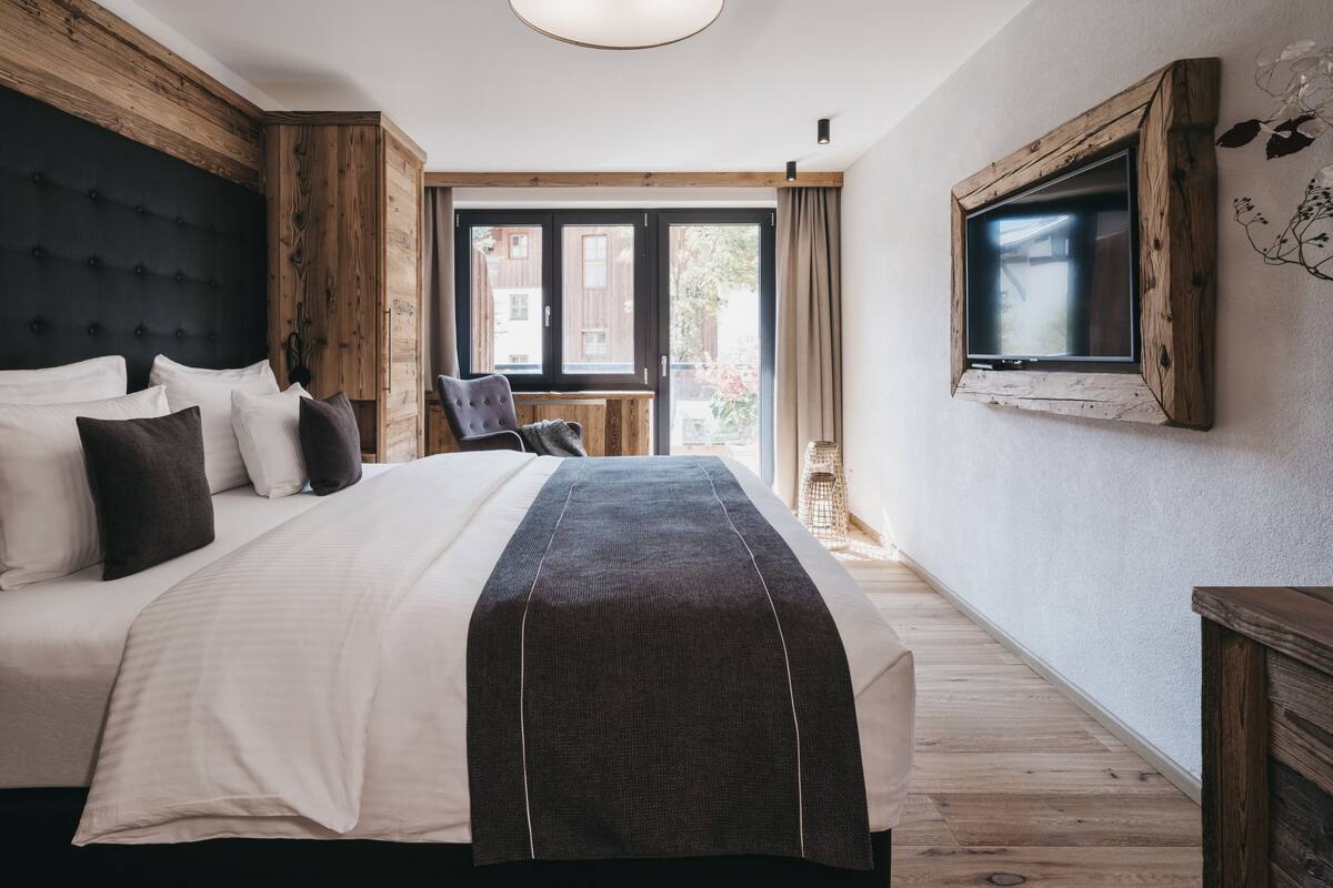 A modern bedroom with a large bed, wooden headboard, a TV mounted on the wall, and a view of the outside through a window.