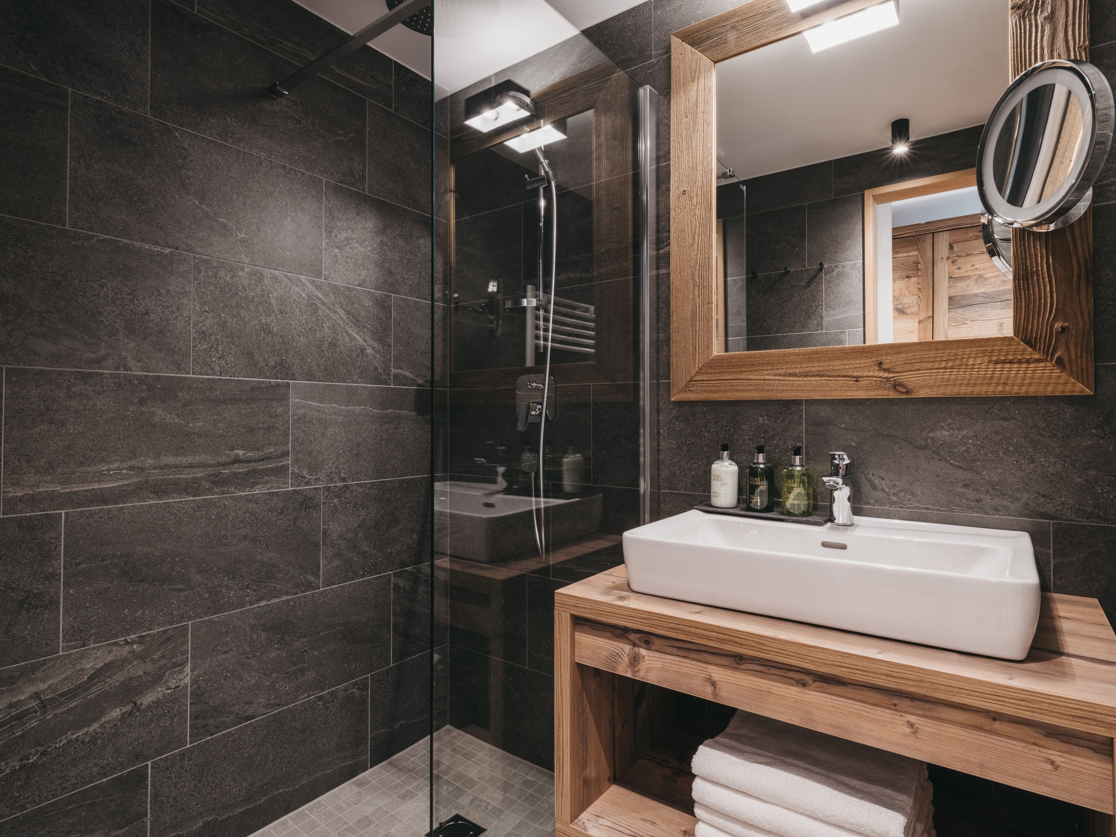 A modern bathroom with dark tiles, a glass shower enclosure, a wooden vanity, and a mirror with a round frame.