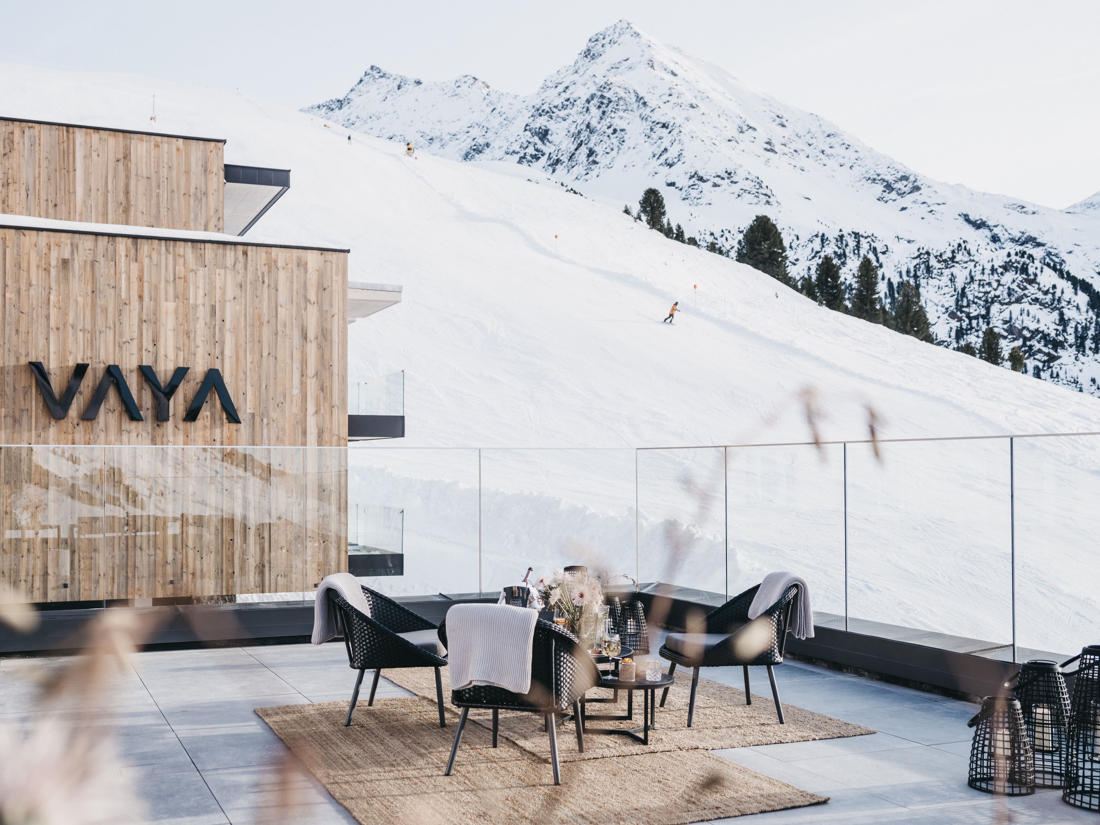 A modern outdoor seating area with a mountain view, a wooden wall with the word 'VAYA' on it, and a ski slope in the background.