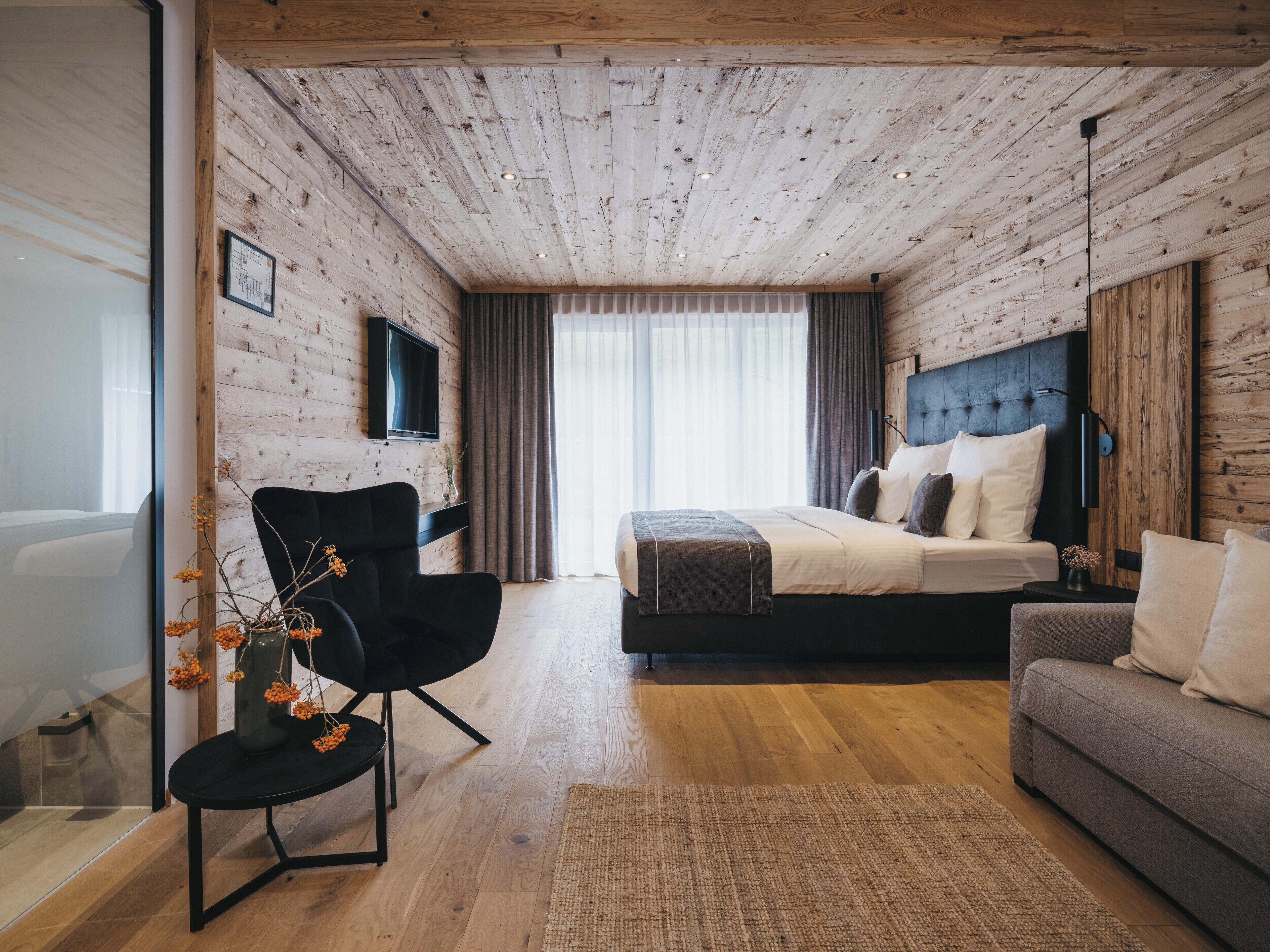 Un dormitorio moderno con techo de madera, una cama grande, un sofá gris y una silla negra.
