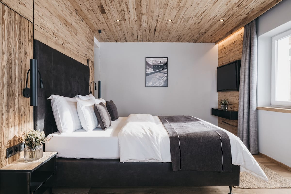 A modern bedroom with a wooden ceiling, a large bed with white and dark bedding, a mounted TV, and a window with curtains.