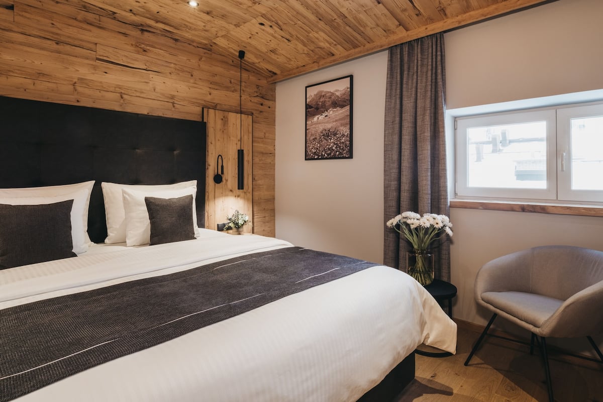 A bedroom with a wooden ceiling, a bed with white and dark bedding, a nightstand with a lamp, a framed picture on the wall, a window with curtains, and a chair.