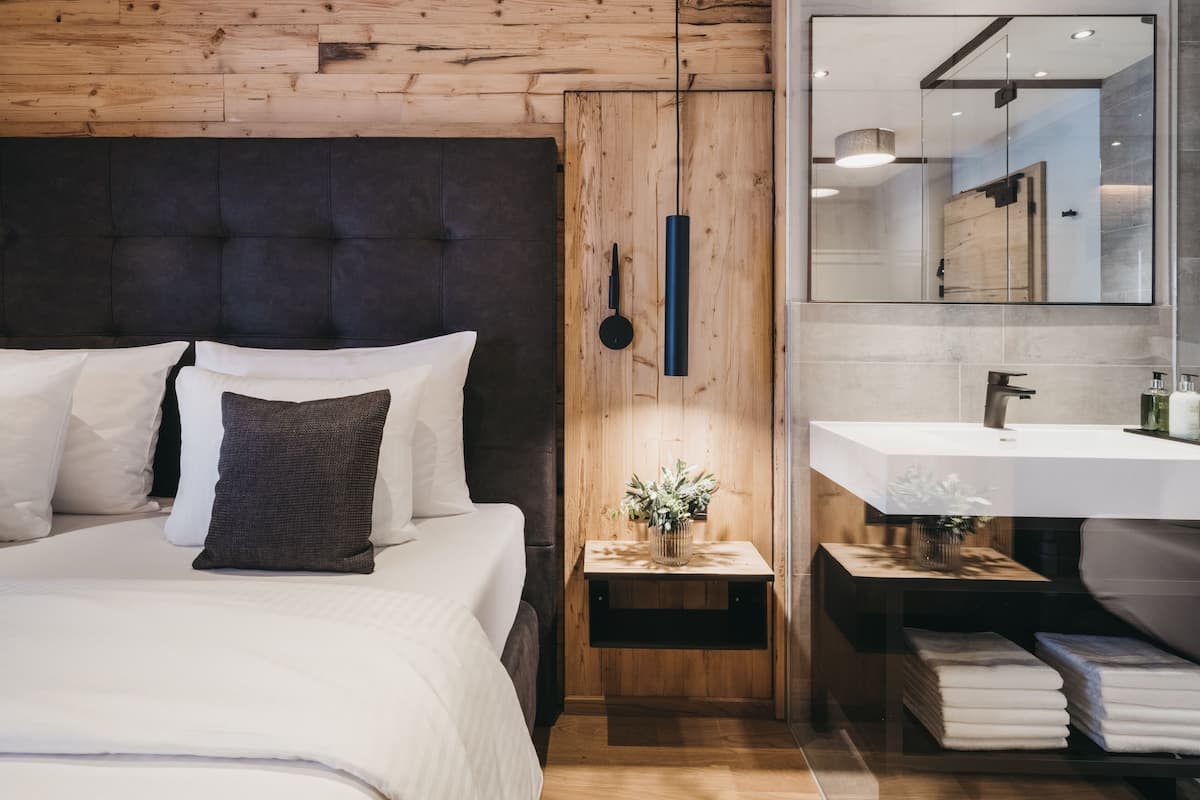 A modern bedroom with a wooden headboard, a bedside table with a vase, and a large mirror above a bathroom vanity.