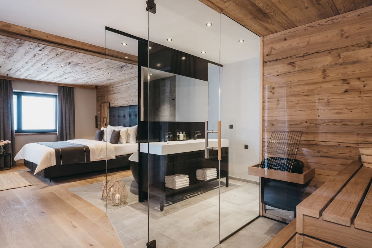 A modern bedroom with a large bed, a glass partition, a wooden bench, and a sauna area.