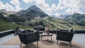 A terrace with two black wicker chairs, a small round table, and a vase with flowers. The terrace overlooks a mountainous landscape with a clear sky. - VAYA Kühtai (Silz)
