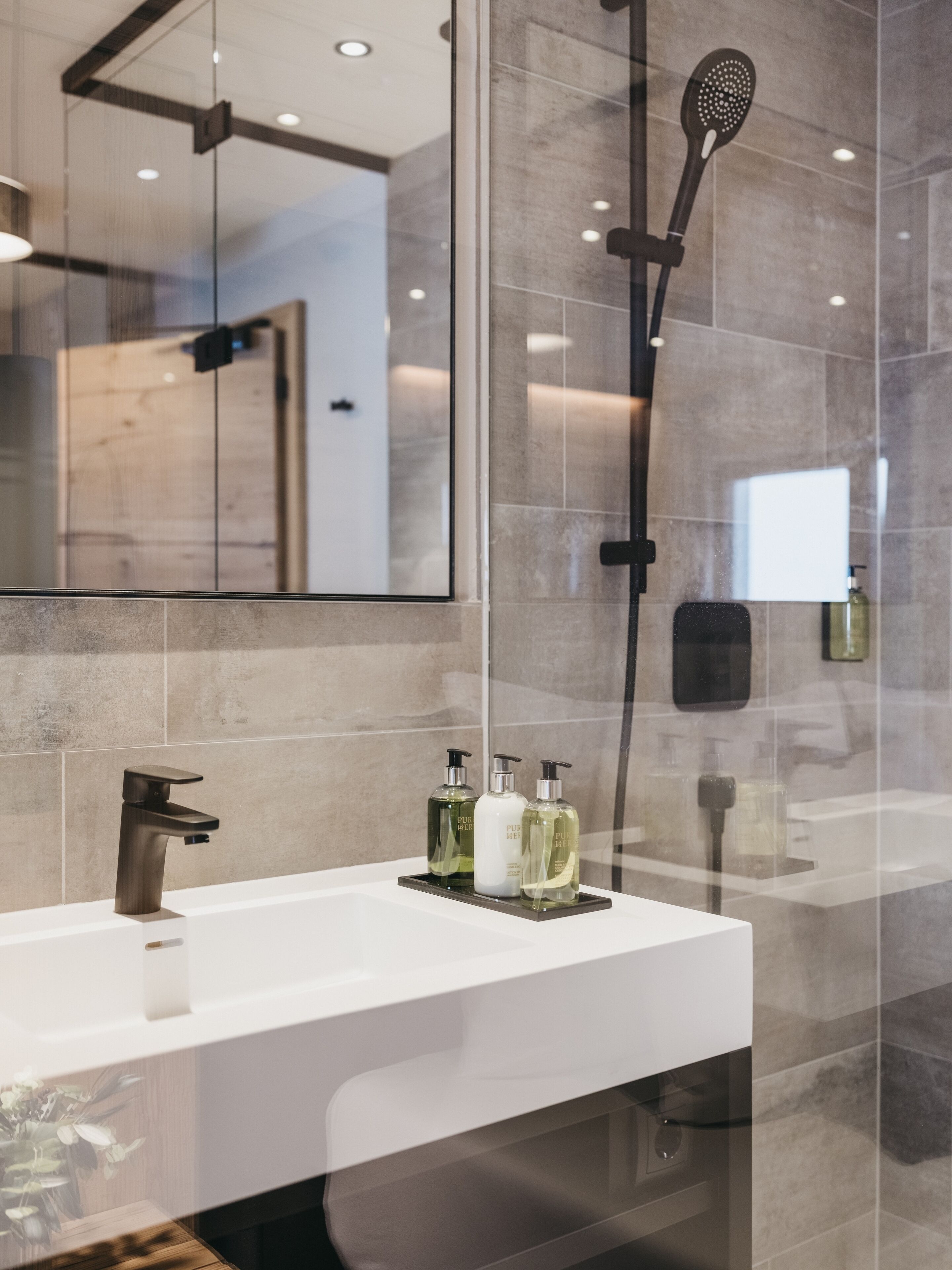 A modern bathroom with a white sink, a handheld showerhead, and three bottles of liquid soap on the counter.