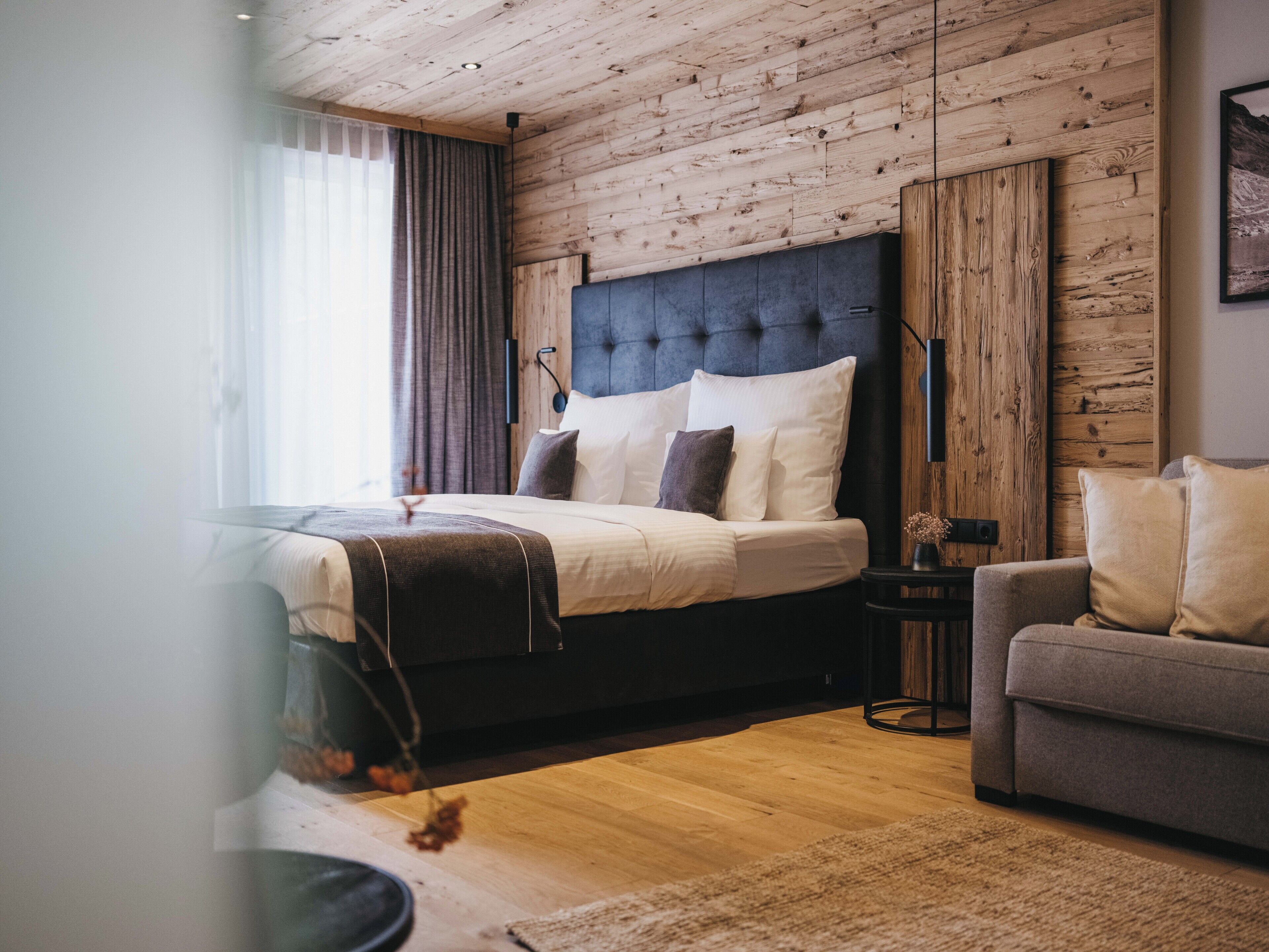 Une chambre moderne avec un plafond en bois, un grand lit avec de nombreux oreillers, un canapé gris et une petite table d’appoint.