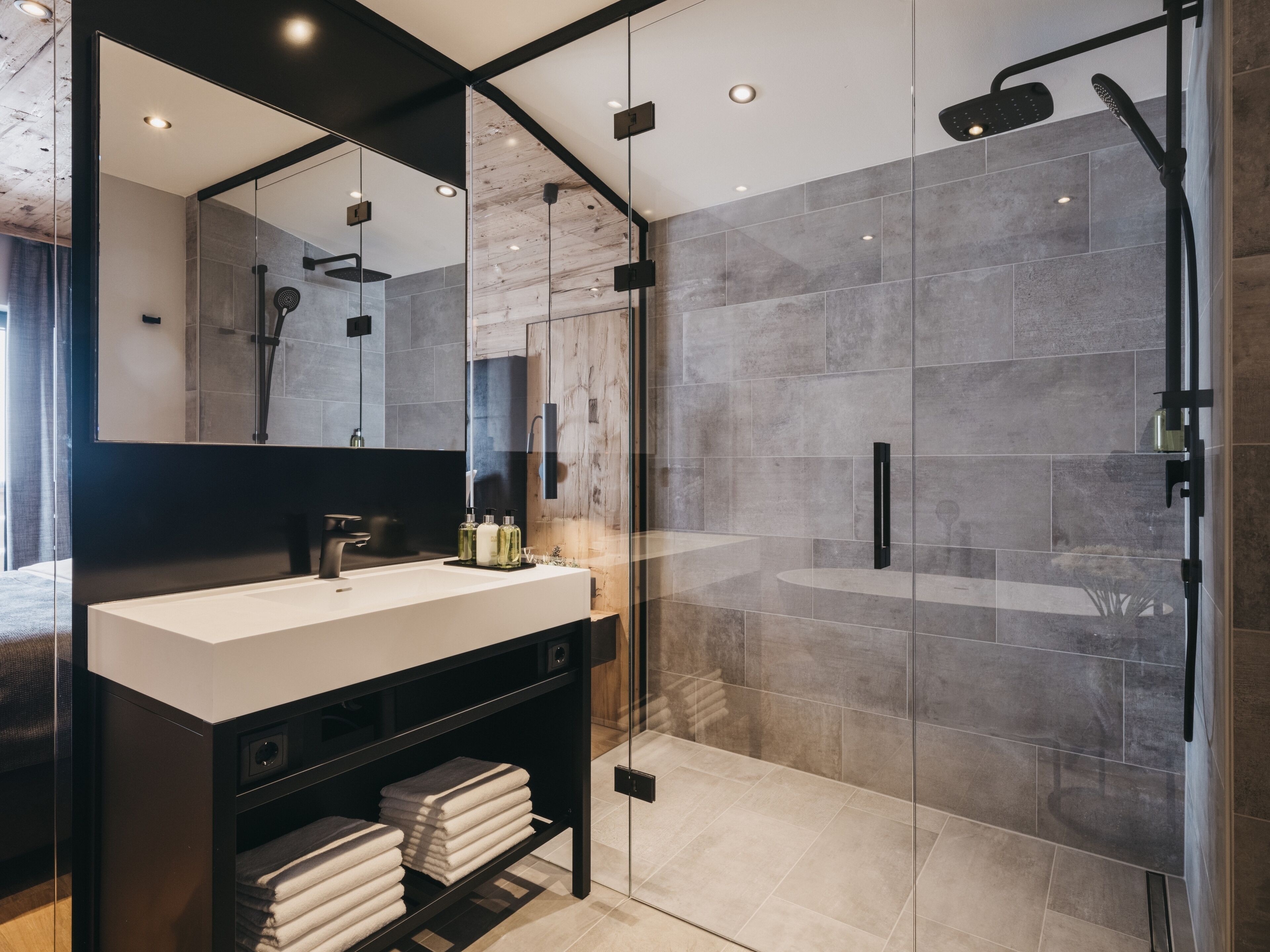 Ein modernes Badezimmer mit einer in Glas abgeschirmten Dusche, einem großen Spiegel und einem weißen Waschbecken mit schwarzem Unterschrank.