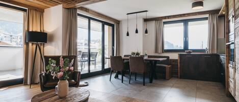 A modern living room with a wooden ceiling, a dining area, and a kitchen. There are large windows providing natural light and a view of the outdoors.
