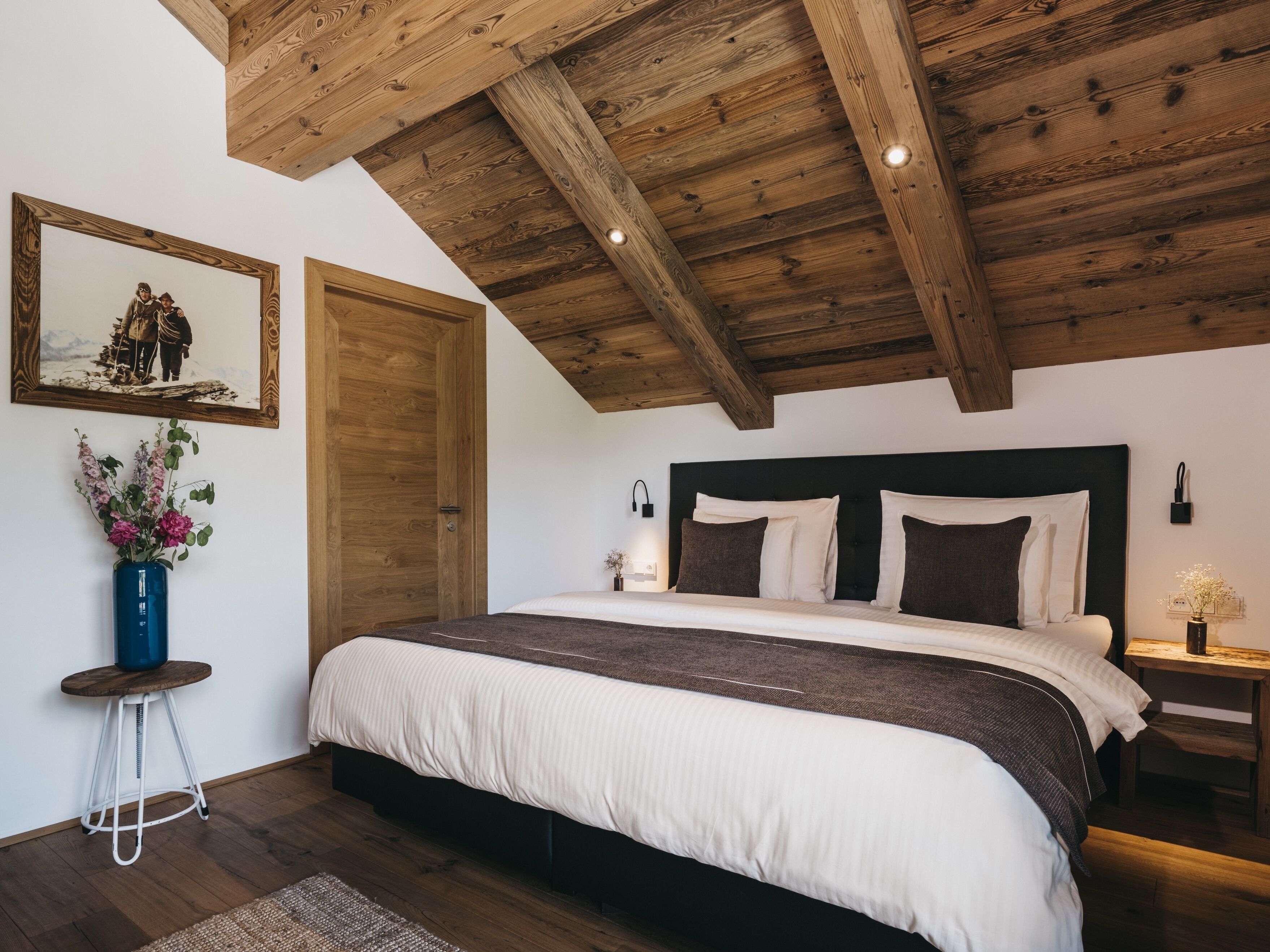 A bedroom with a wooden ceiling, a large bed with white and dark bedding, a wooden door, and a framed picture on the wall.