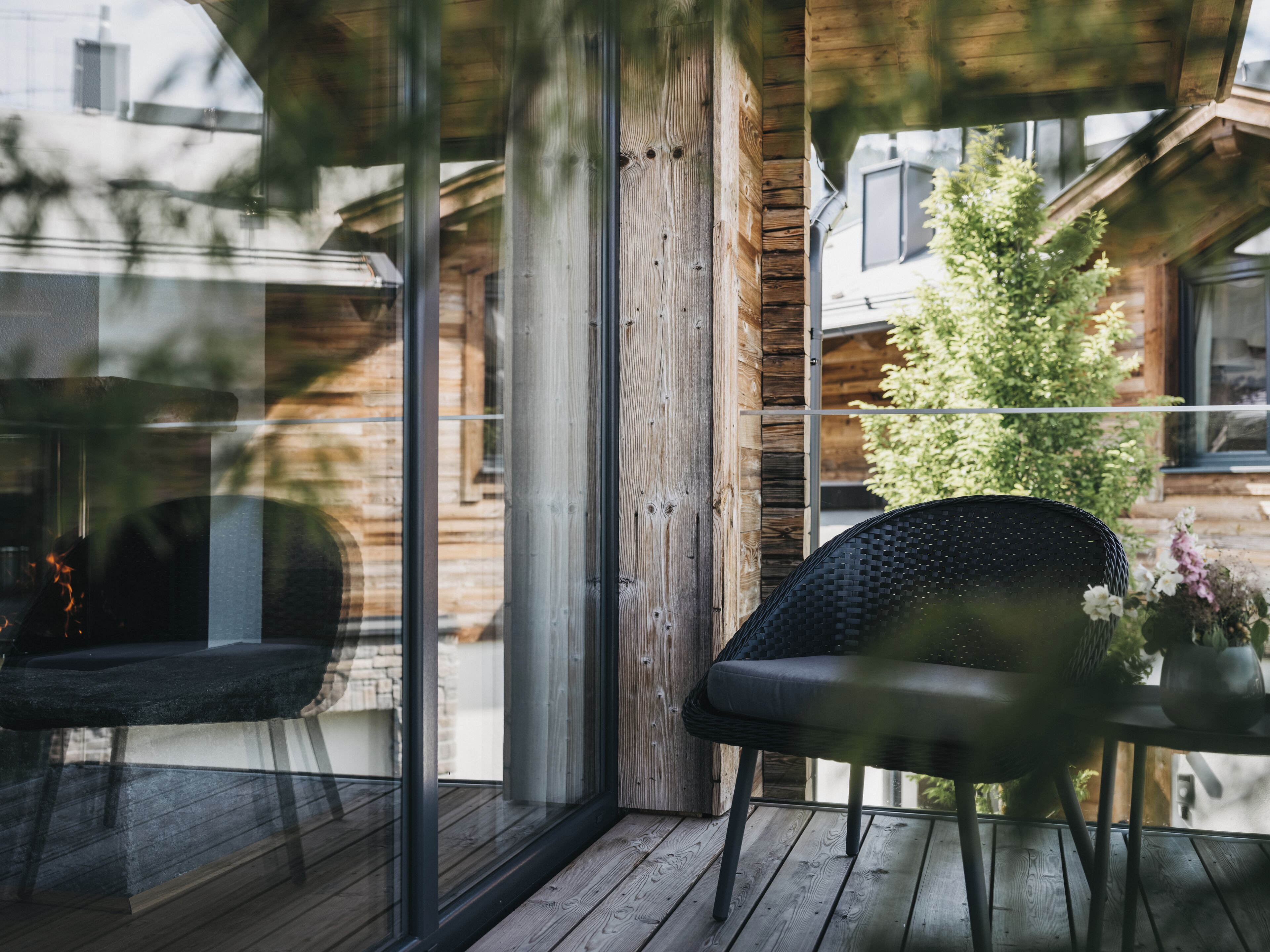 Eine Holzterrasse mit einer Glastür, die auf einen Balkon führt, ein Korbstuhl und ein kleiner Tisch mit einer Blumenvase.