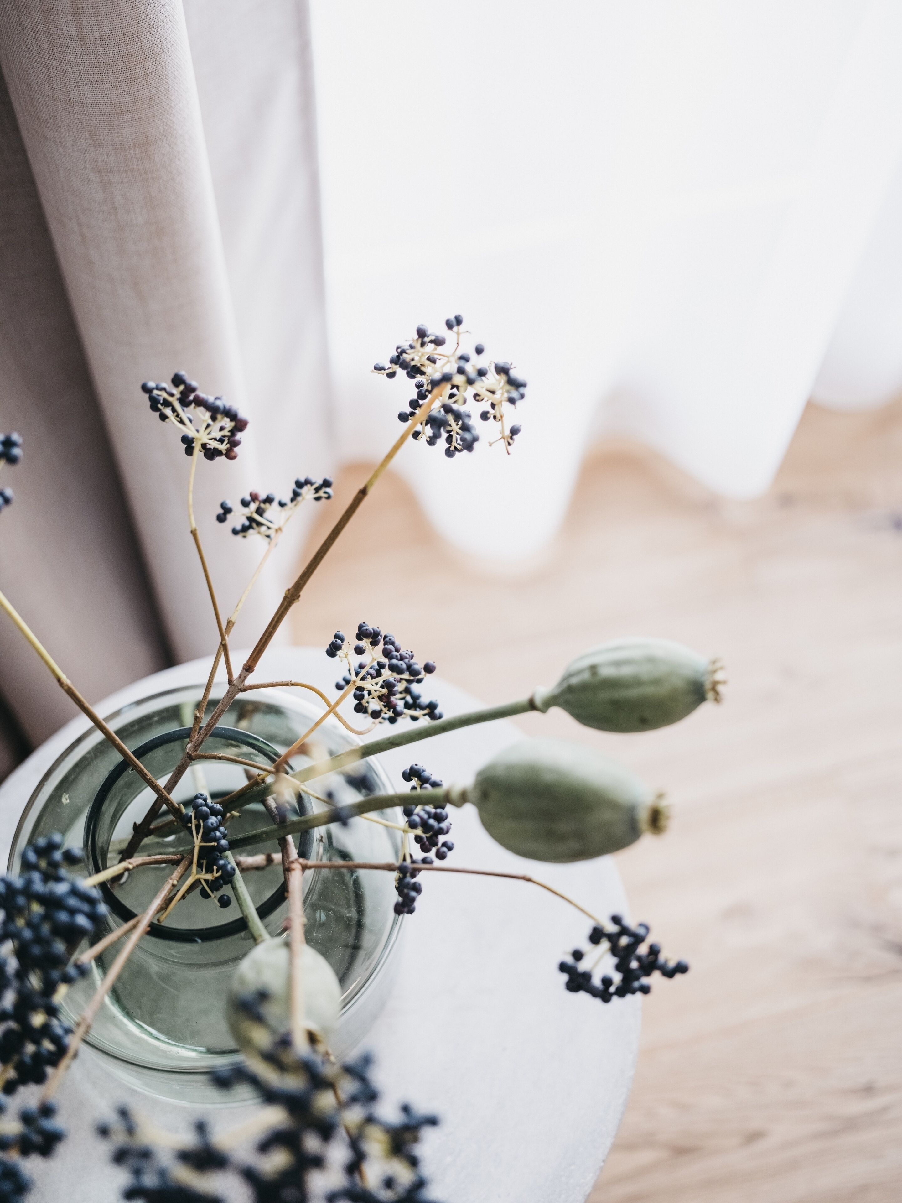 A clear glass vase with dried flowers and green buds on a wooden surface.