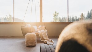 A room with a large window, a hanging chair, a woven ottoman, and a bench.