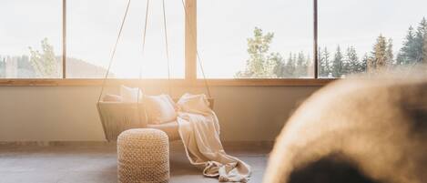 A room with a large window, a hanging chair, a woven ottoman, and a bench.