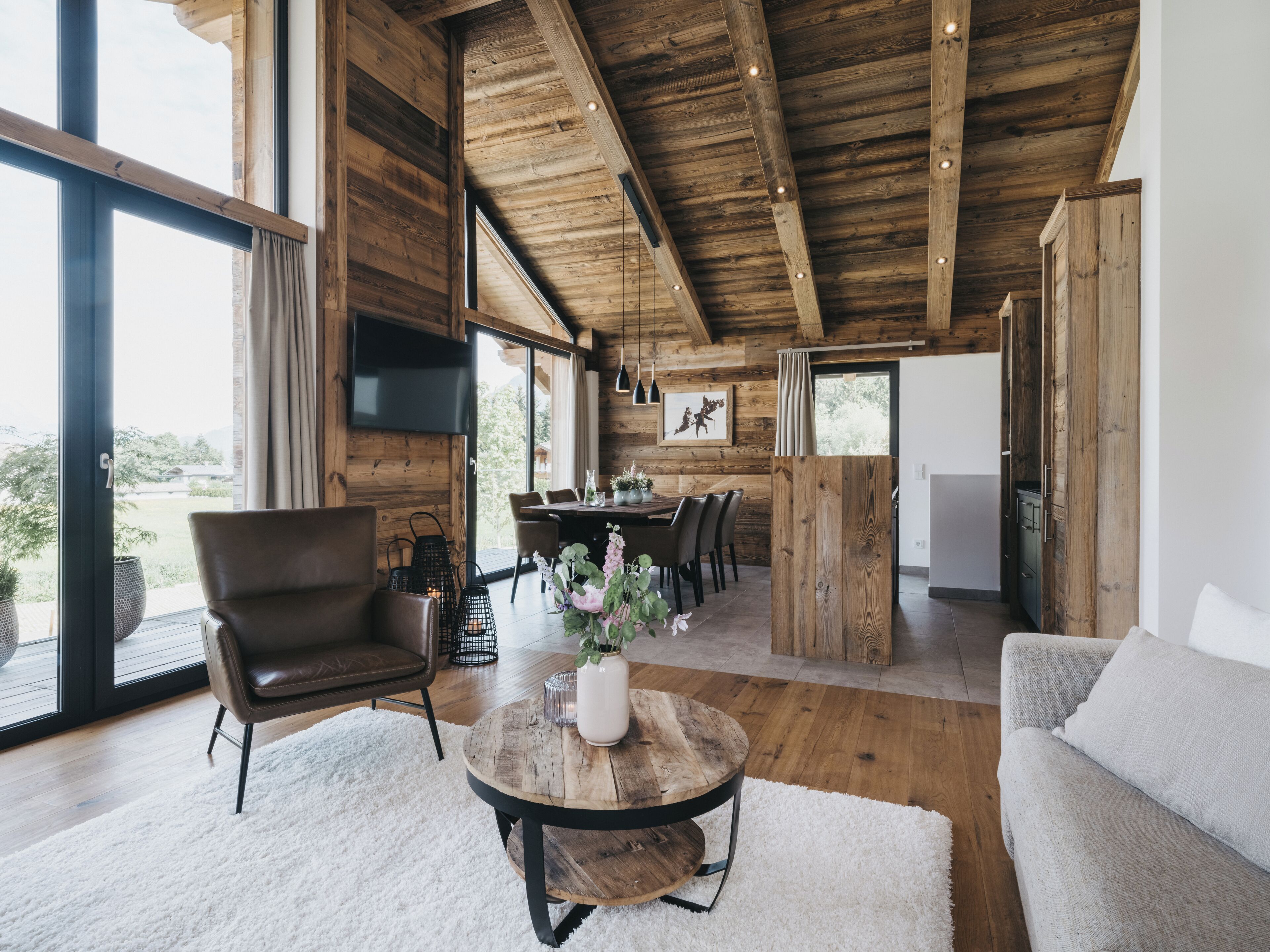 A spacious living area with a wooden ceiling, a dining table set for six, and a large window offering a view of the outdoors.