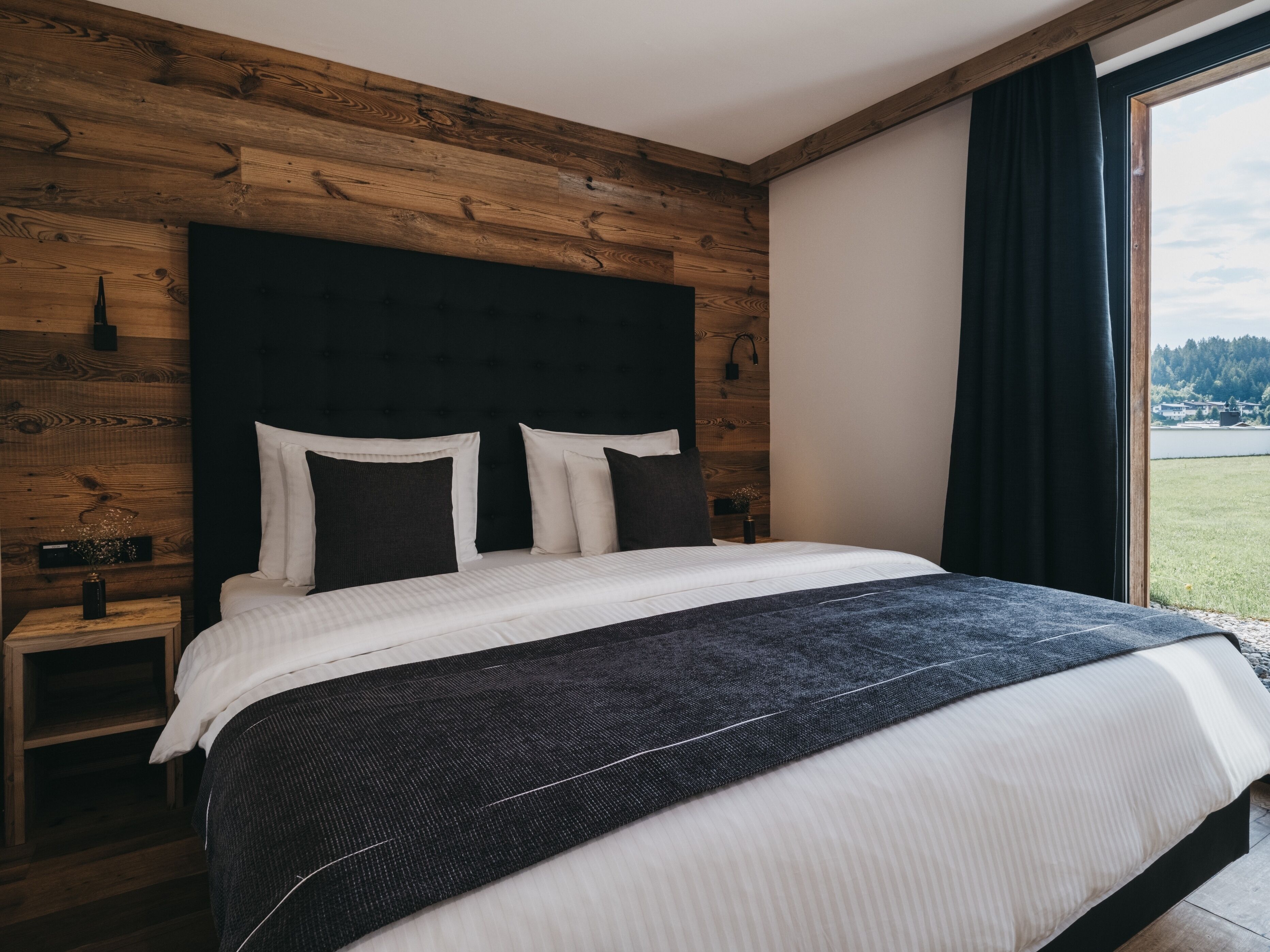 A bedroom with a wooden headboard, a bed with white and dark bedding, a nightstand with a vase, and a view of a green landscape through a window.