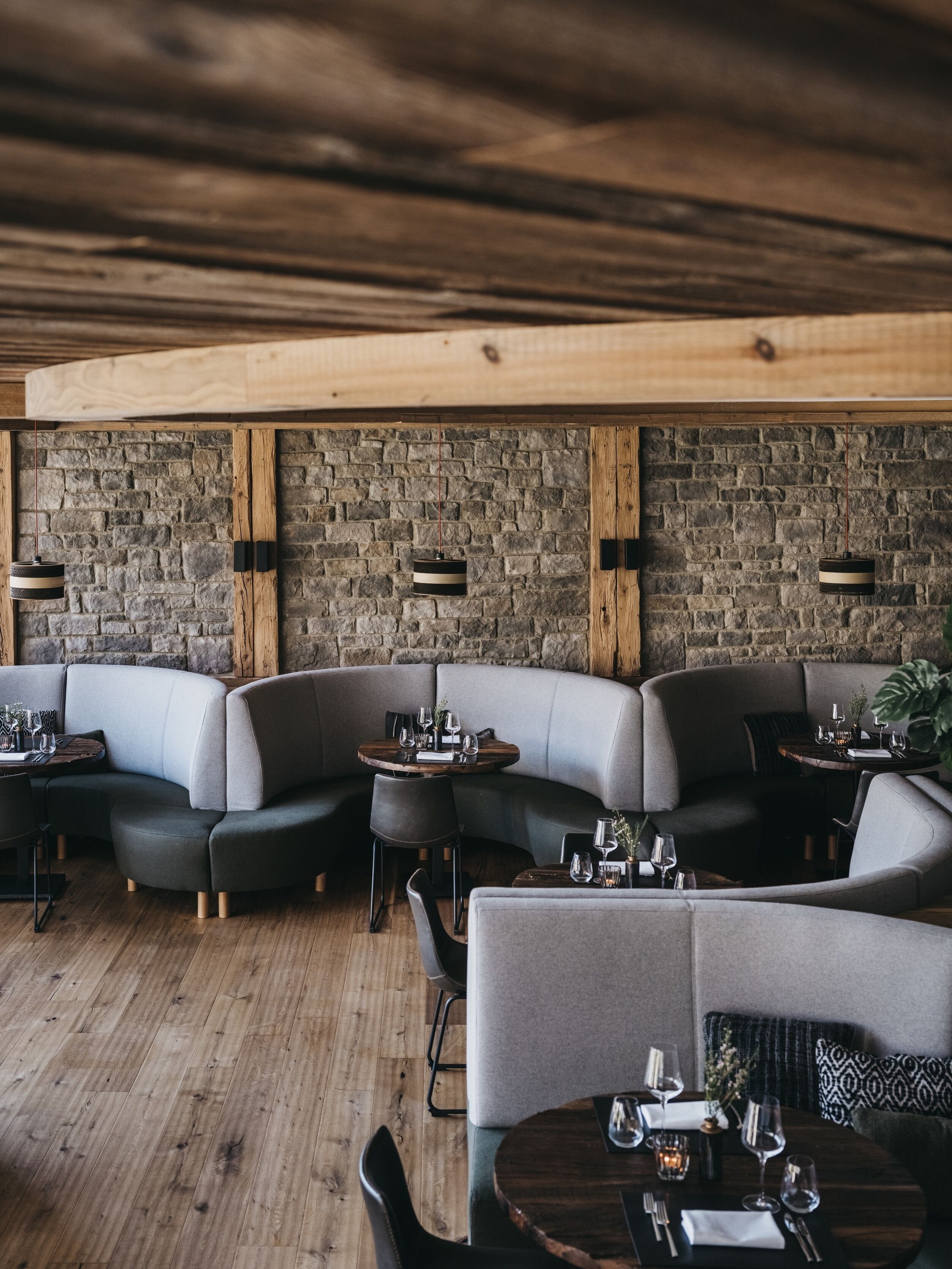 A modern restaurant with a stone wall, wooden ceiling beams, and grey upholstered seating.