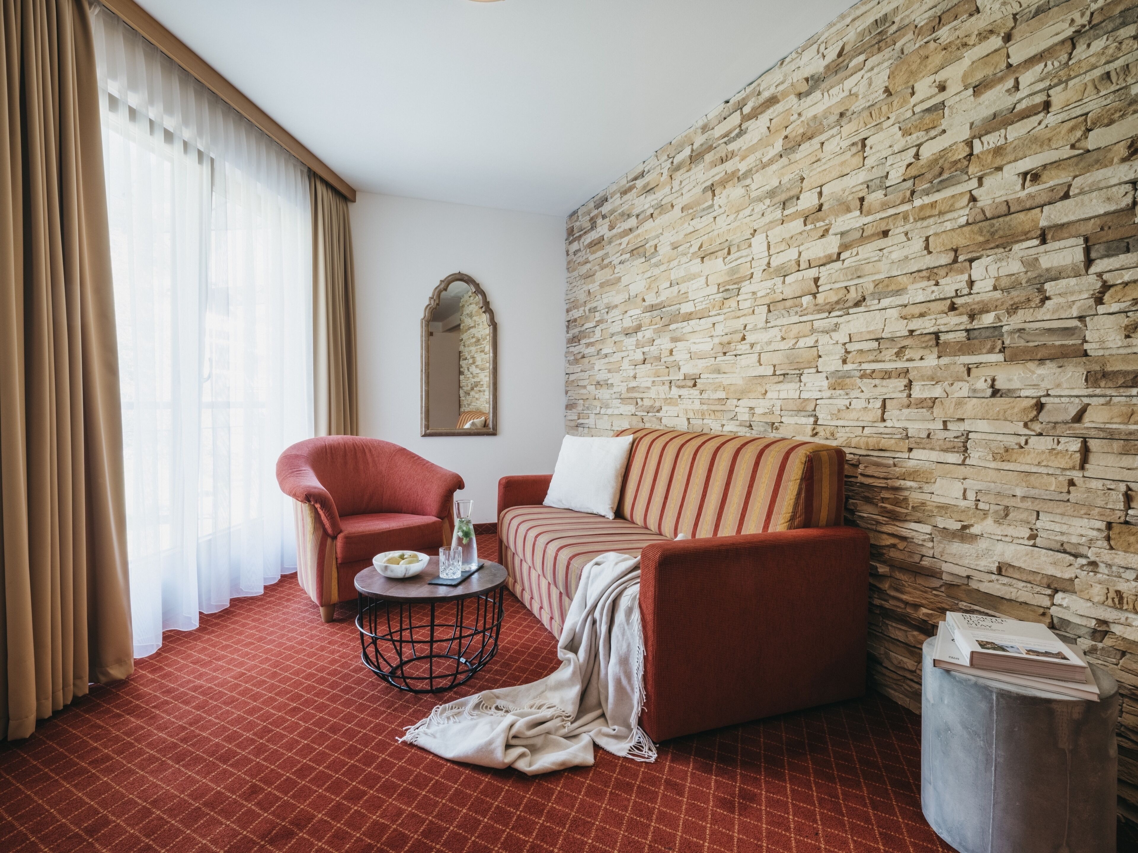 A room with a stone wall, a red sofa, a red armchair, a round table with a vase, and a mirror on the wall.