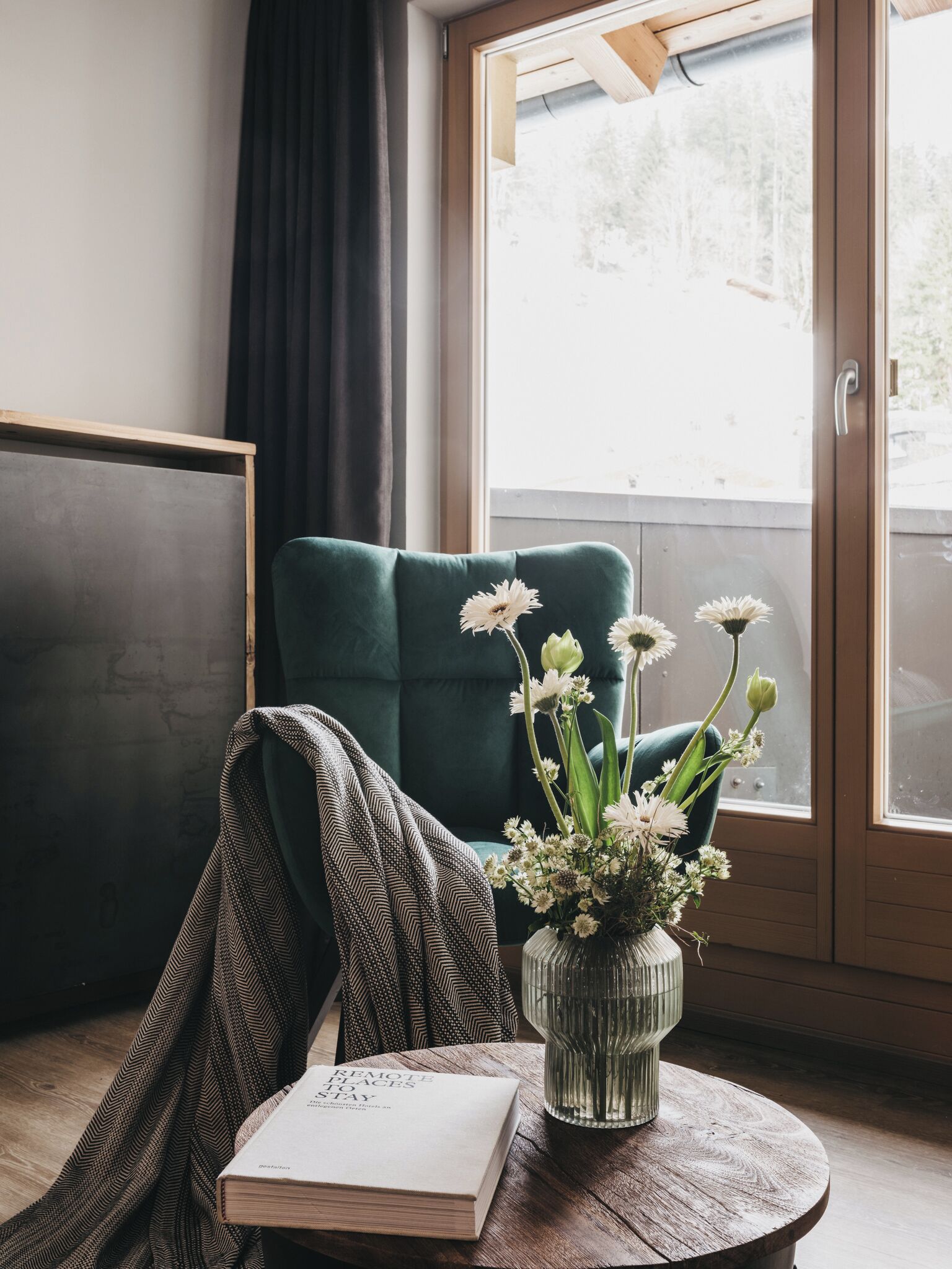 A room with a teal chair, a wooden table, a book titled "Remote Places to Stay," and a vase with flowers.