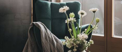 A room with a teal chair, a wooden table, a book titled "Remote Places to Stay," and a vase with flowers.