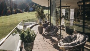 Een modern balkon buiten, met glazen hekwerk, planten in potten, rieten stoelen en uitzicht op een groen veld en bergen.