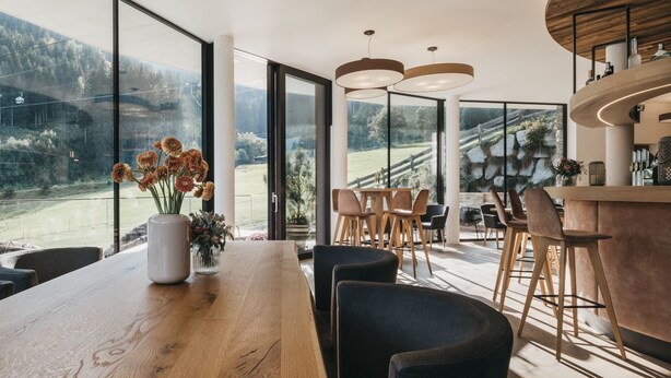 Een moderne keuken met grote ramen, een houten eettafel, barstoelen en een bloemenvaas.