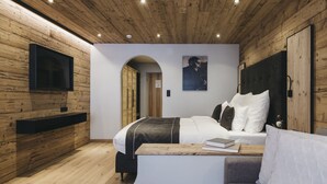 Een moderne slaapkamer met een houten plafond, een flatscreen televisie aan de muur, een bed met wit en grijs beddengoed, een nachtkastje met een boek, en een houten bankje.