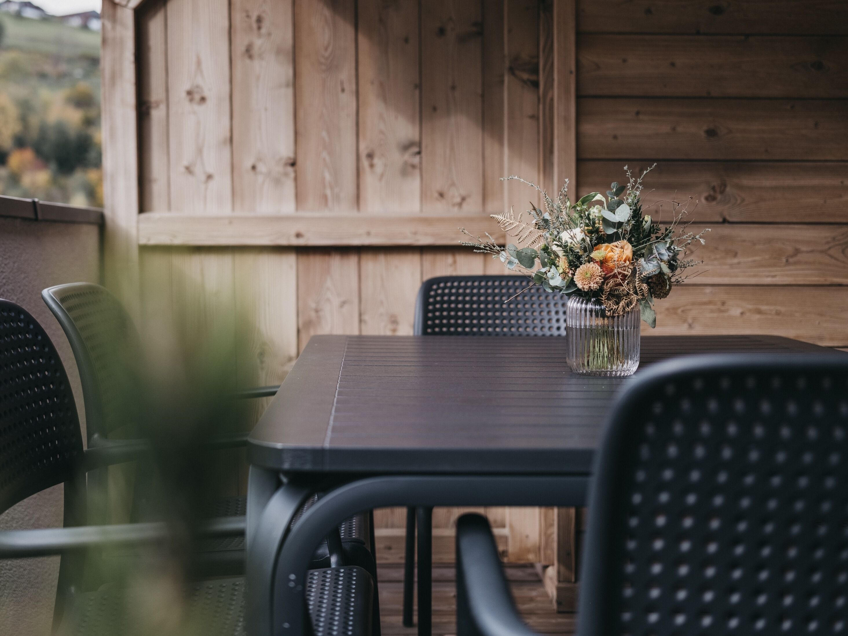 Een houten terras met een tafel, stoelen, een vaas met bloemen en een uitzicht op een houten gebouw achterin.