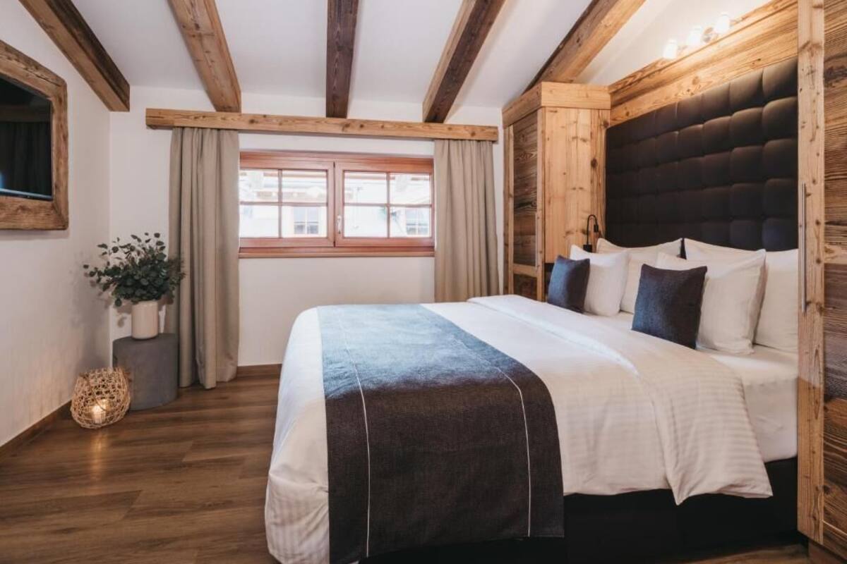 A bedroom with a wooden ceiling, a large bed with a dark headboard, a window with curtains, and a potted plant.
