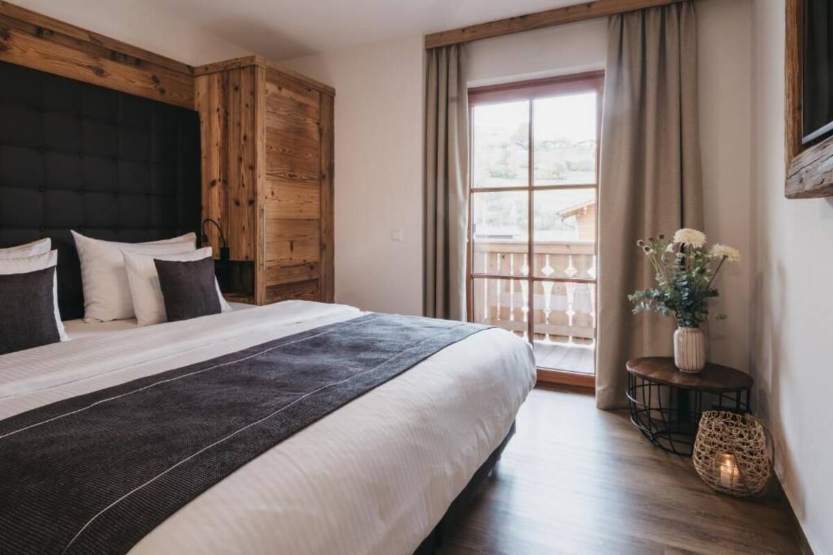 A bedroom with a large bed, wooden headboard, a window with curtains, and a small table with a vase and candle.