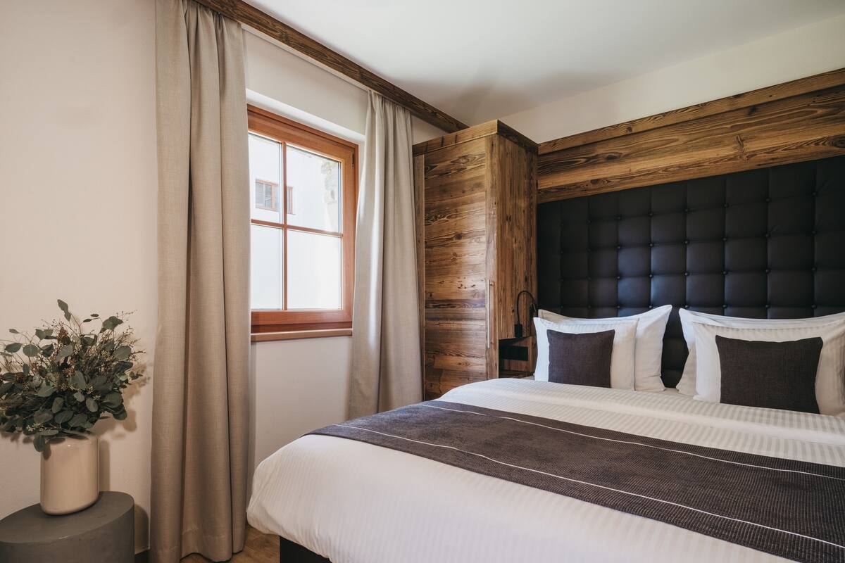 A bedroom with a large bed, wooden paneling, a window with curtains, and a potted plant.
