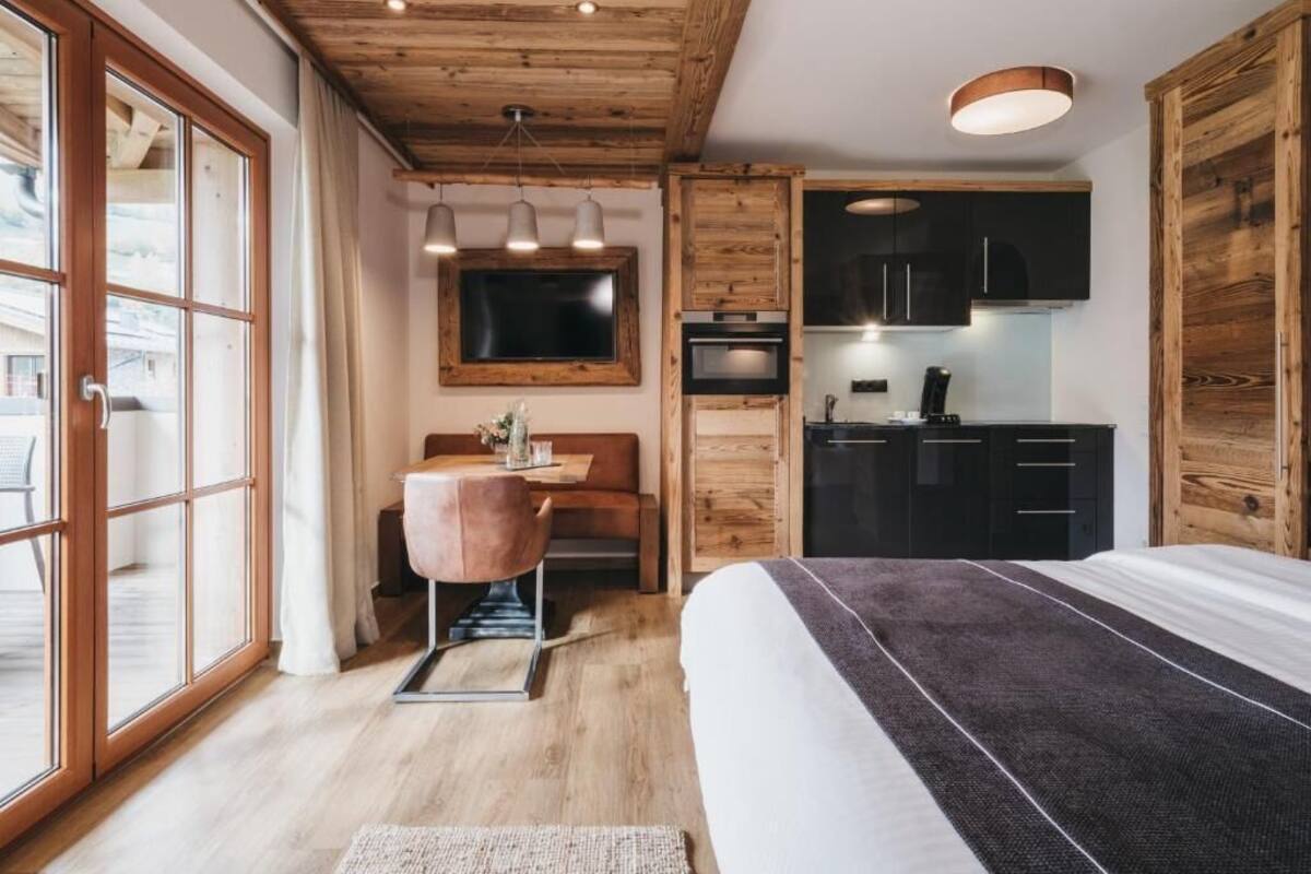 A bedroom with a large bed, wooden ceiling, a desk with a chair, a TV, and a kitchenette with cabinets and a stove.