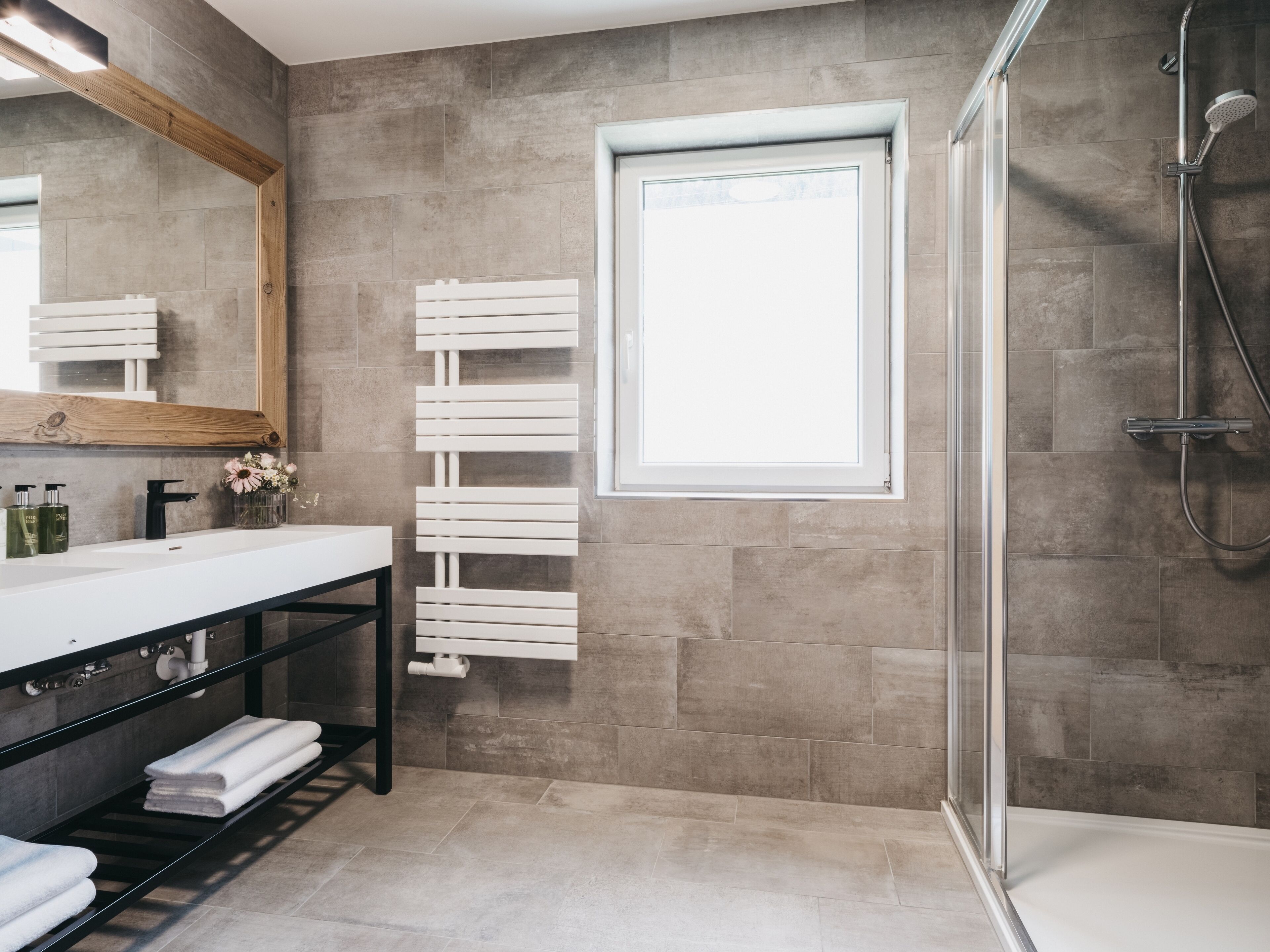 A modern bathroom with a large mirror, a white towel rack, a shower area, and a window.