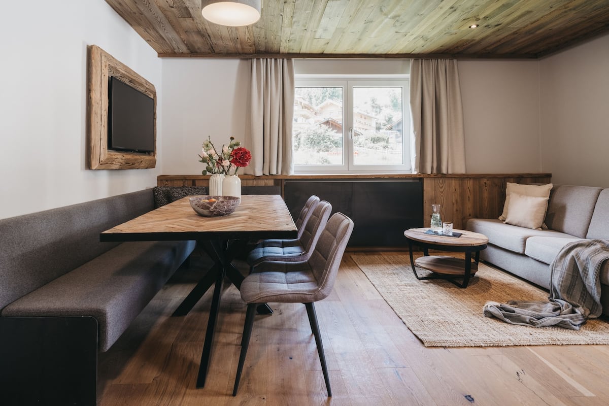 A cozy room with a wooden dining table, bench seating, a sofa, and a window with curtains.